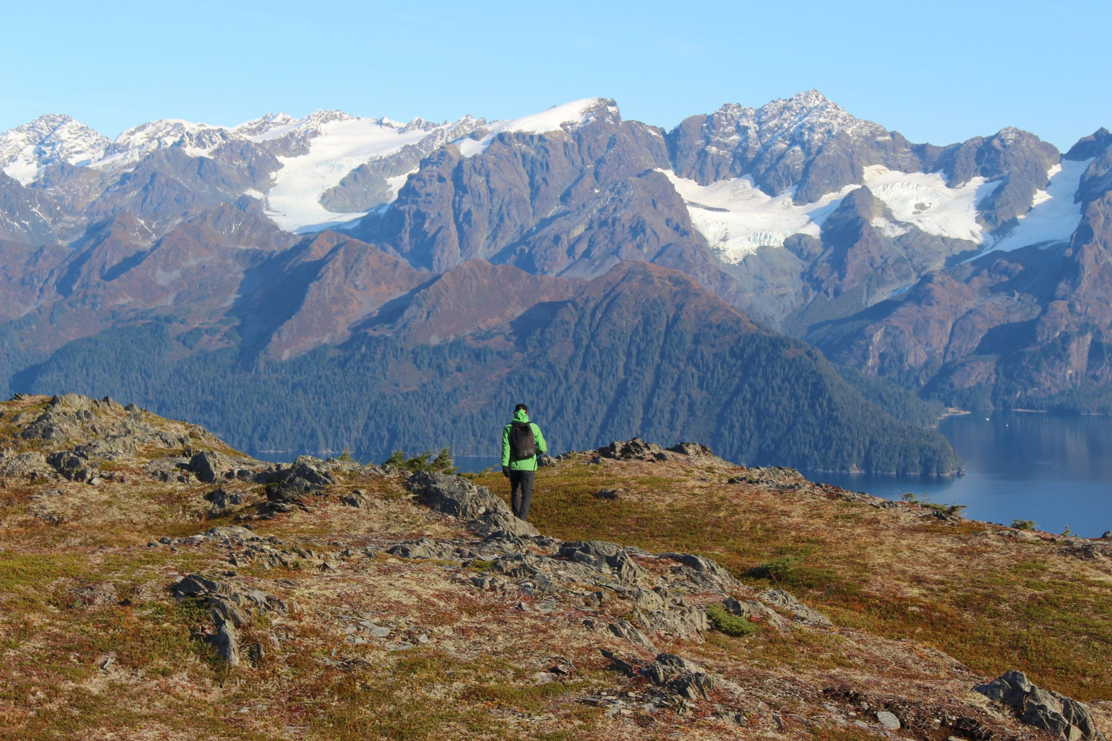 Hiker in green jacket walking on rocky terrain with mountains and water in the background.