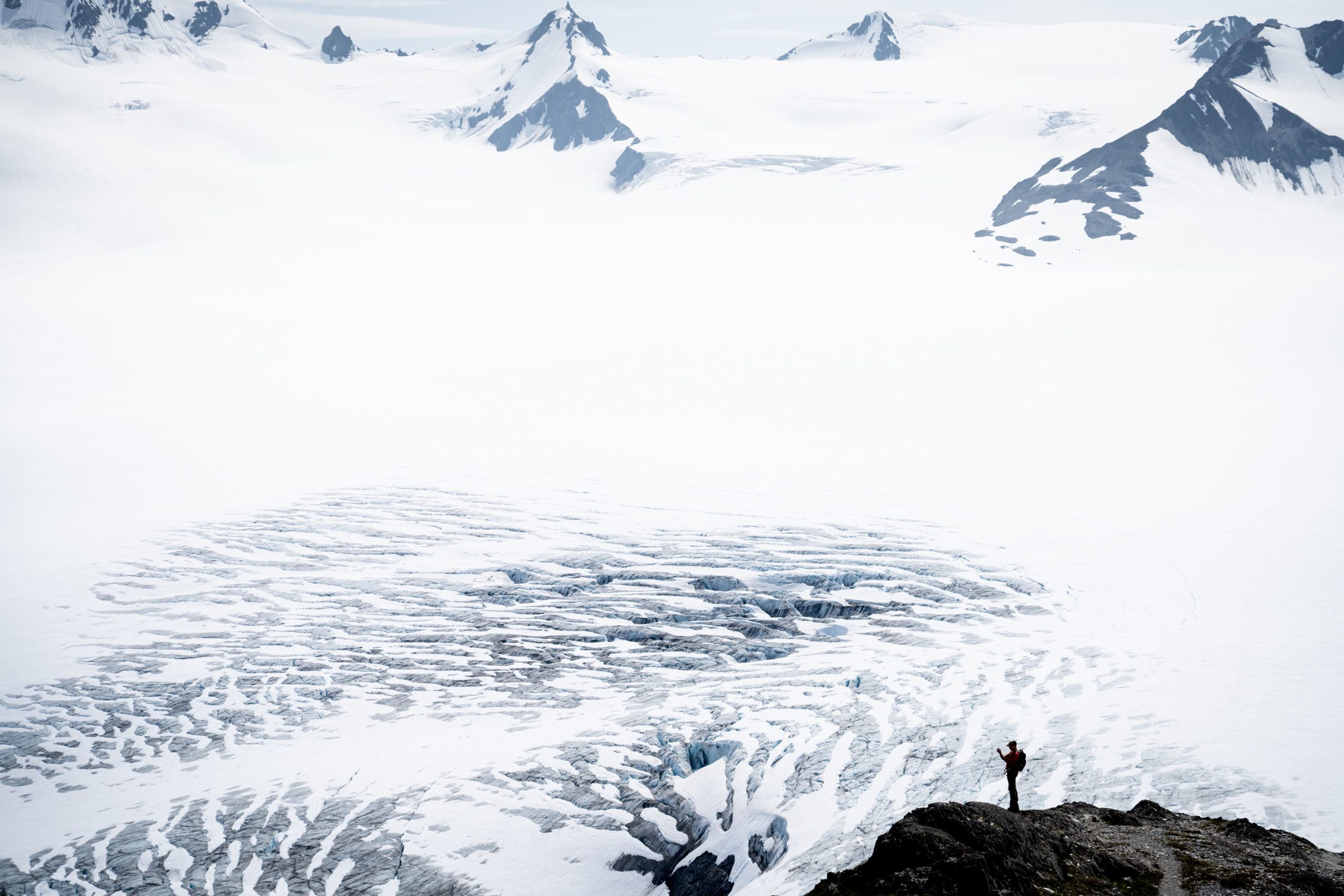 A person stands on a rocky outcrop overlooking a vast glacier landscape.