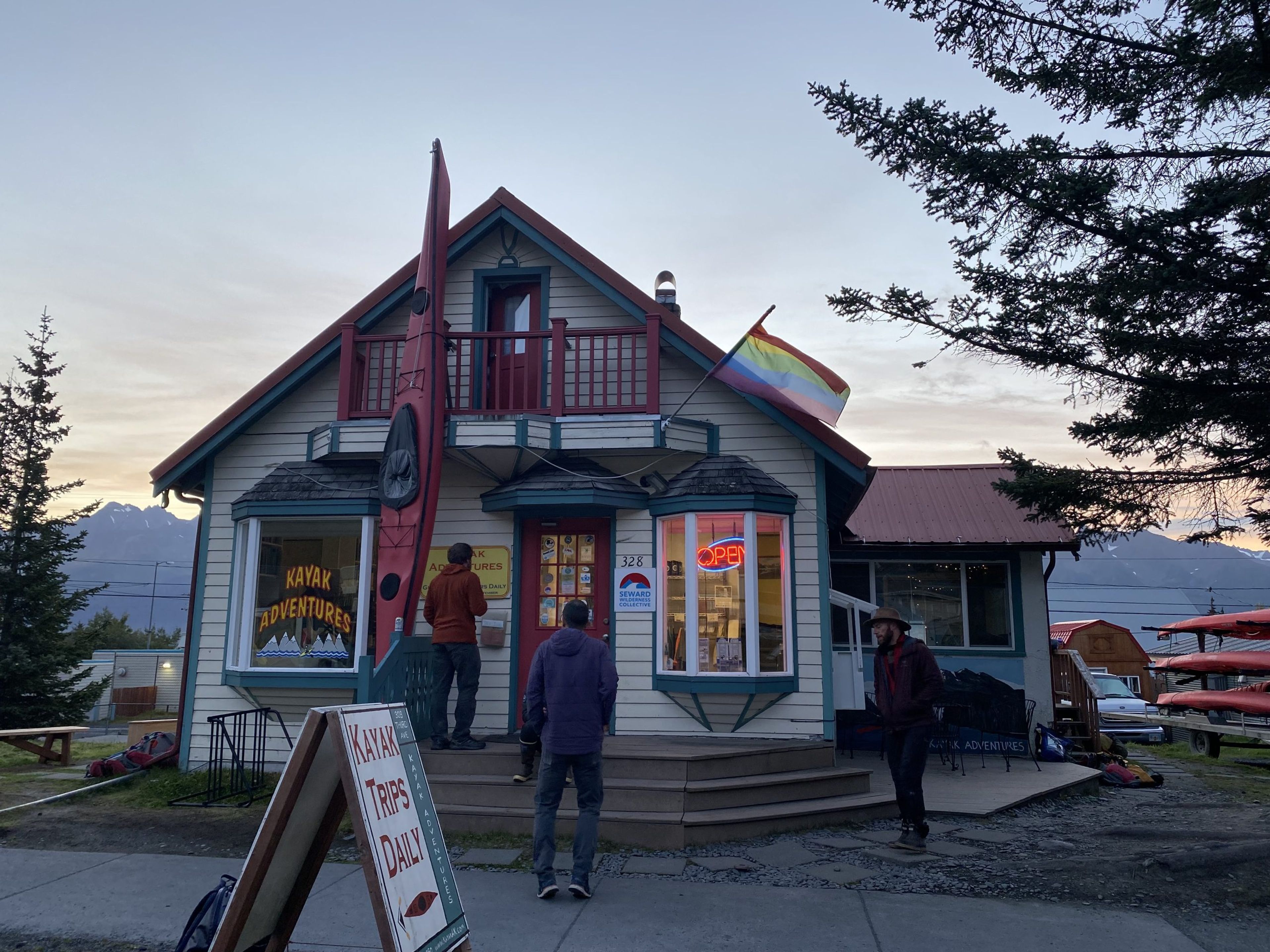 A kayak adventure shop with a rainbow flag and kayaks outside at sunset.