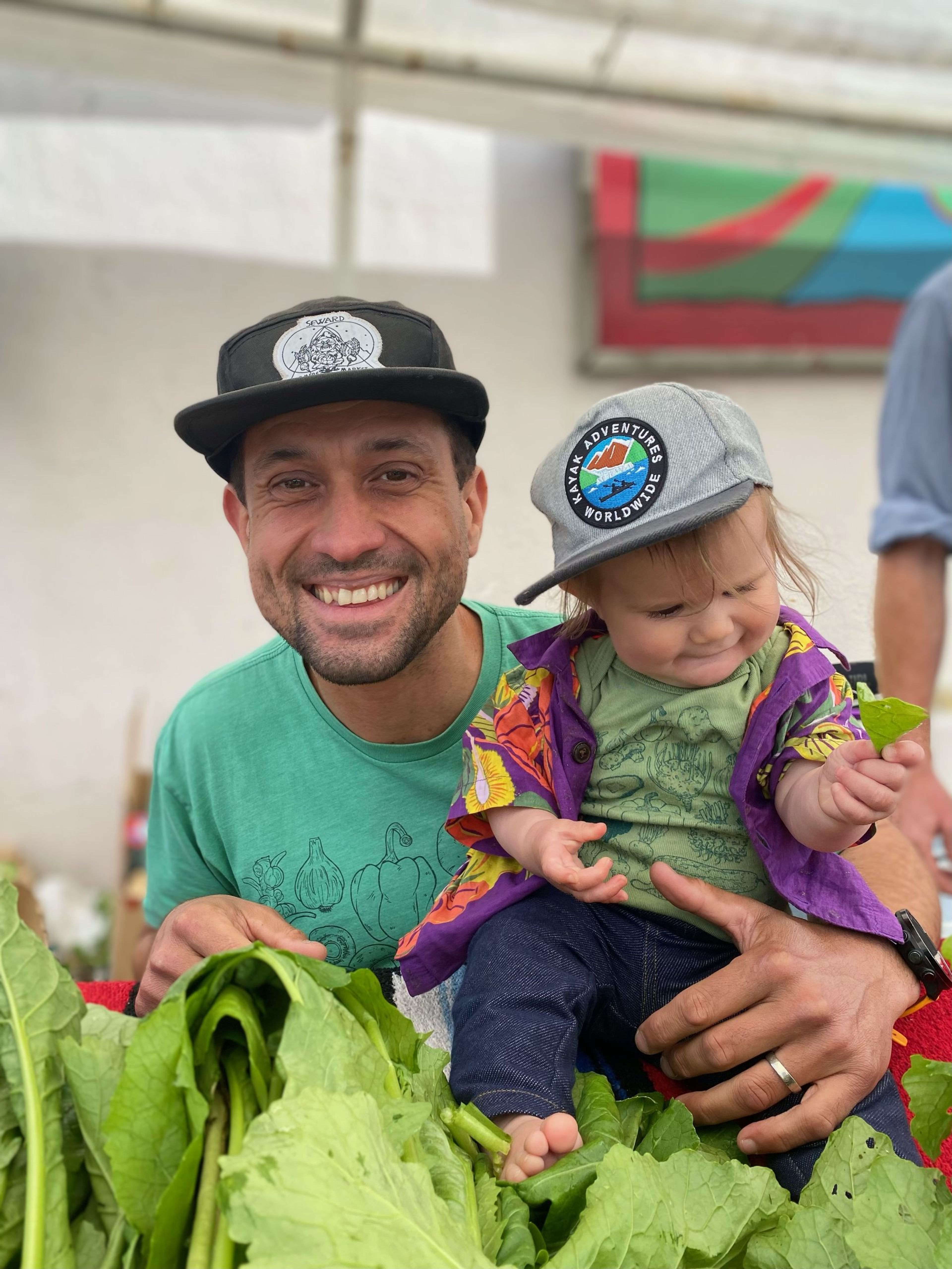A man and a child smiling among fresh greens at a market.