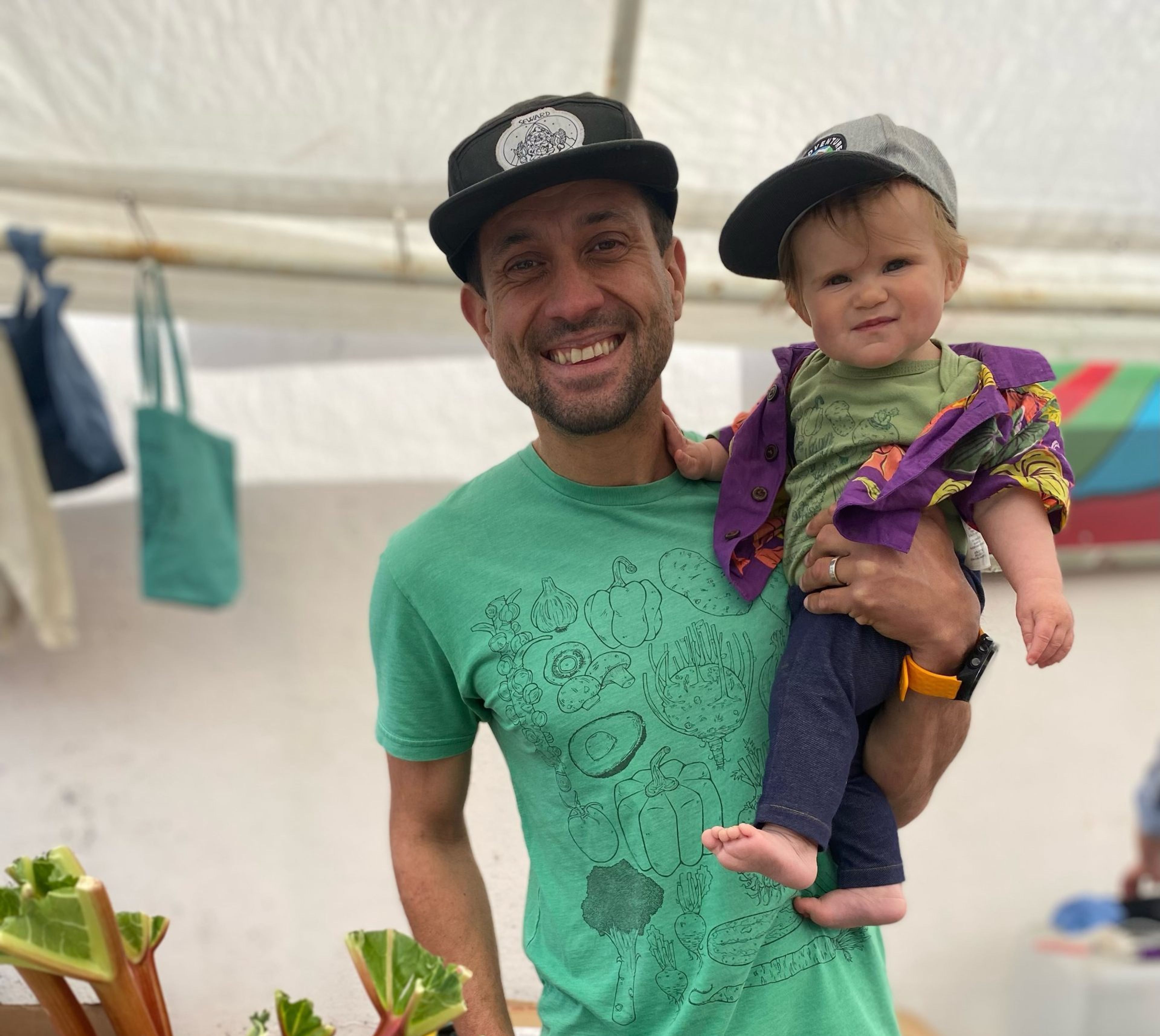 A man holds a baby at a market, both smiling, with fresh produce in the foreground.