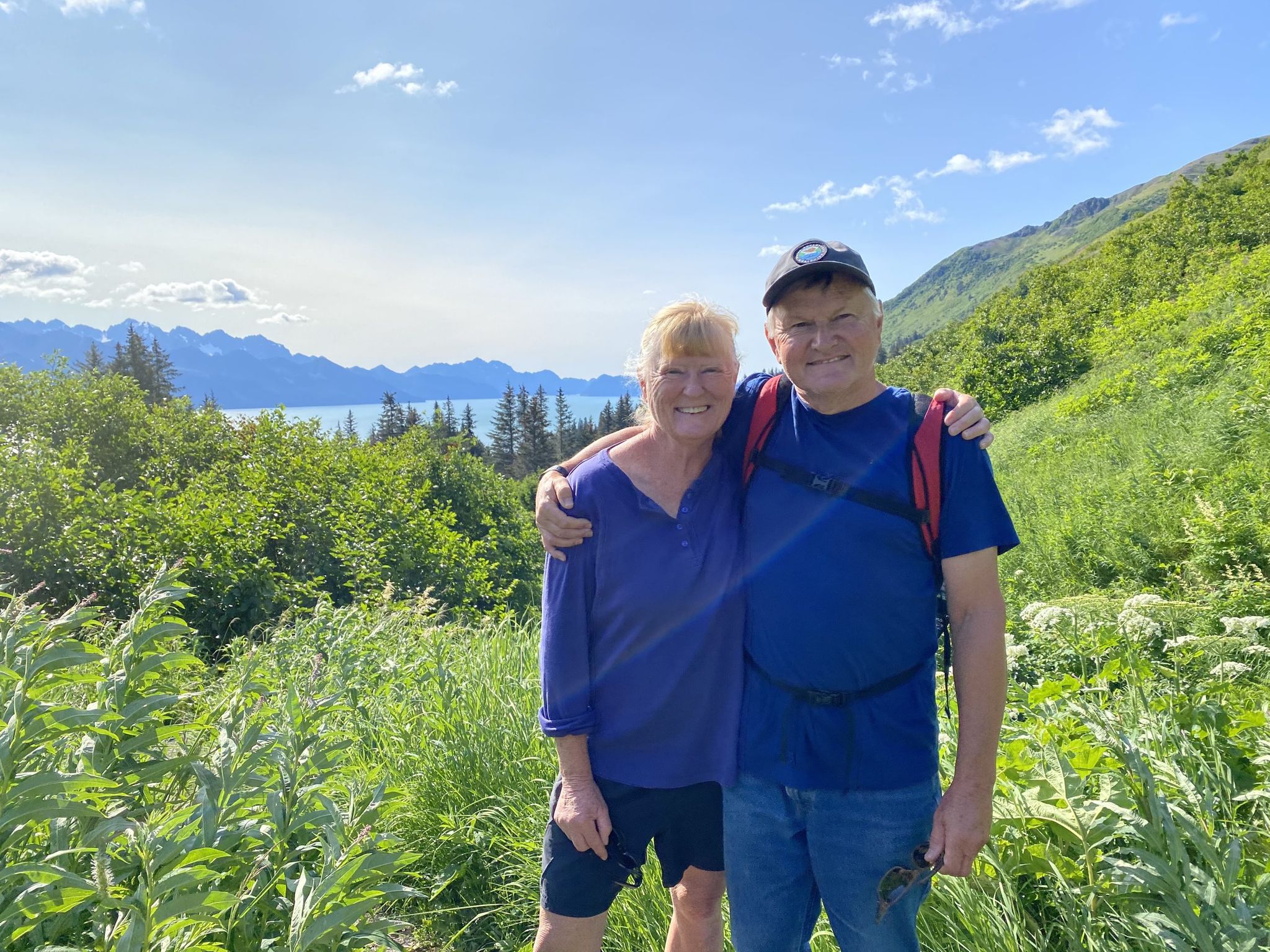 Two people posing together in a lush green landscape with mountains and water in the background.