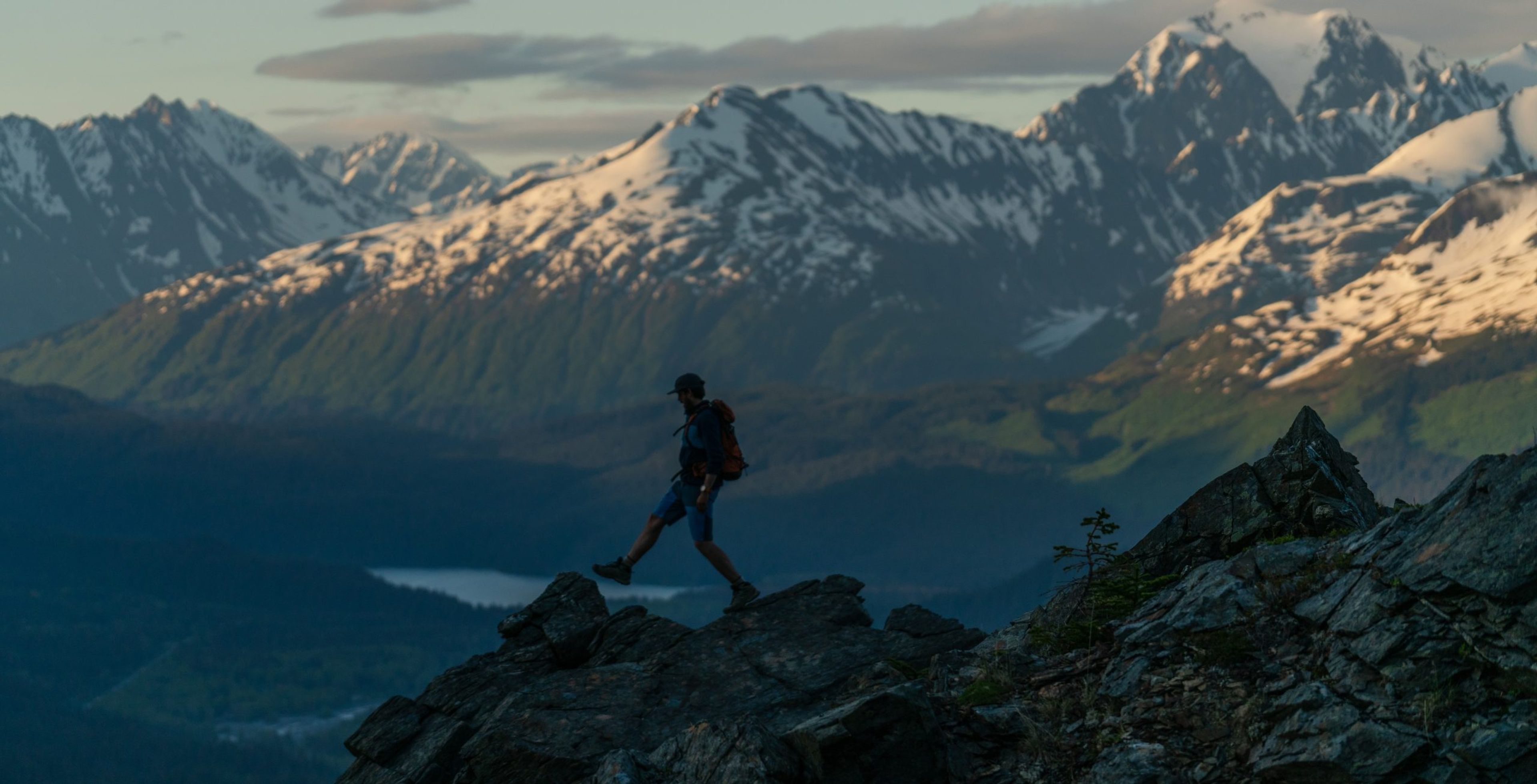 Hiker navigating rocky terrain with mountains in the background during sunset.