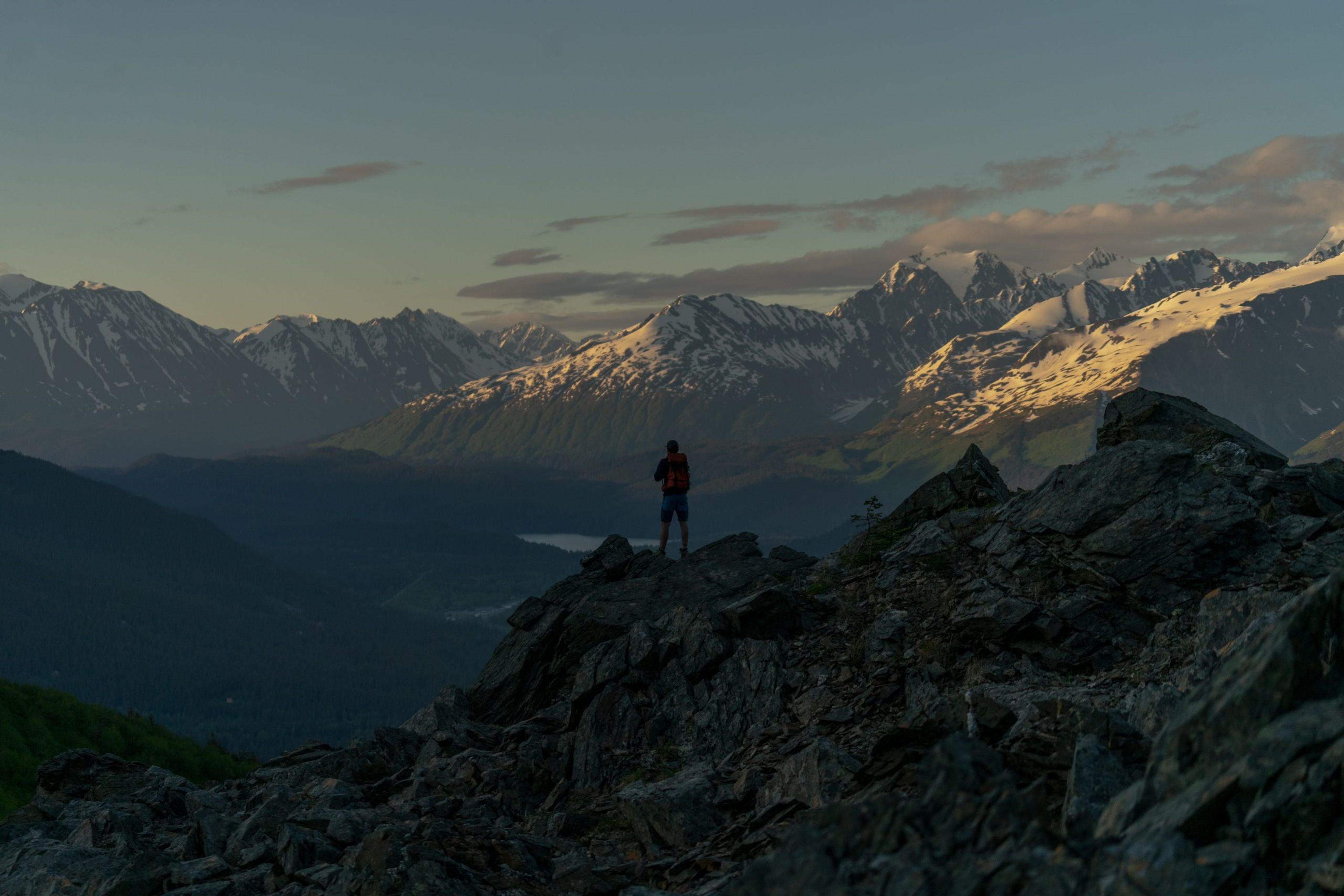 A hiker stands on rocky terrain overlooking snow-capped mountains at sunset.