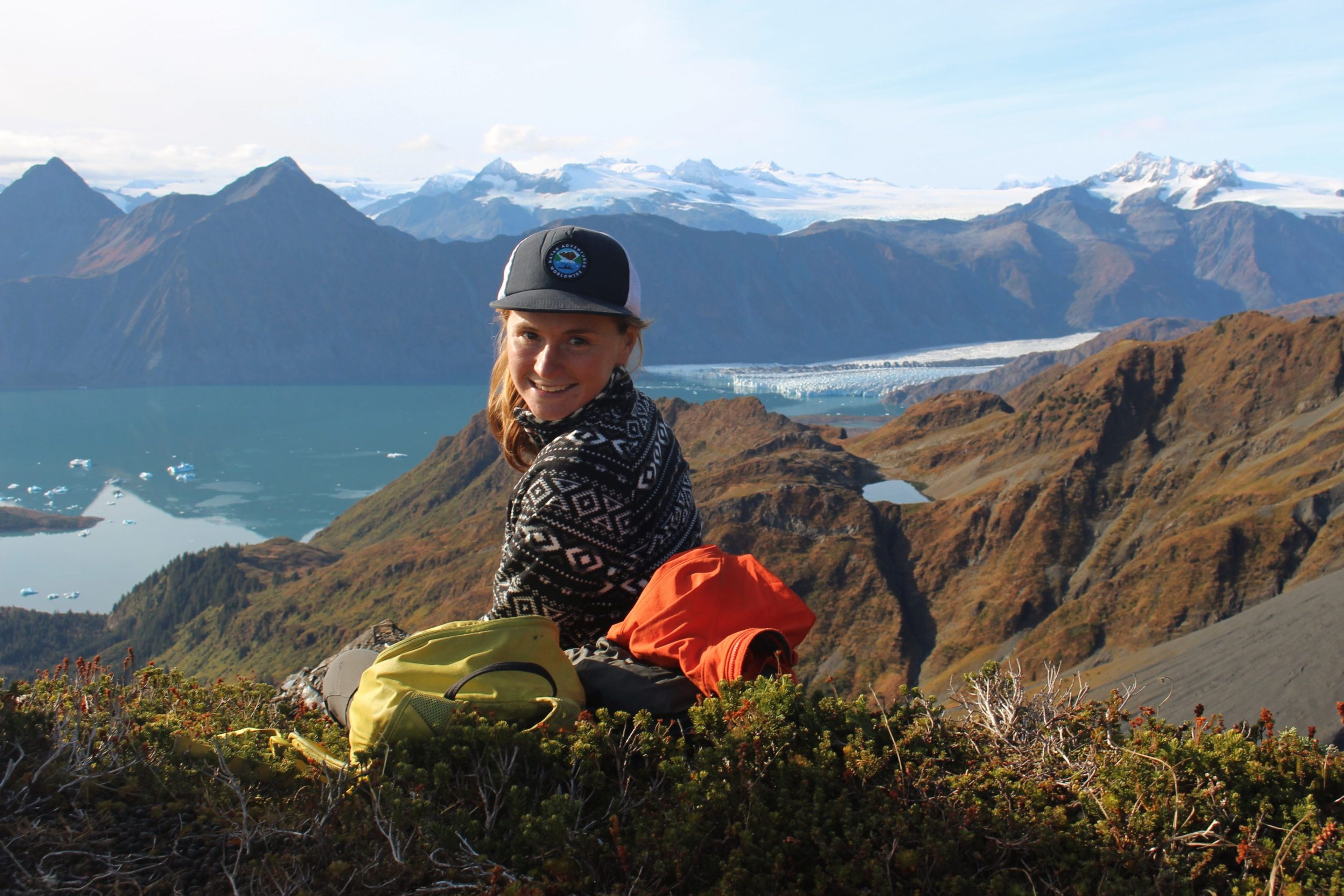 Person sitting on a mountain overlooking a glacier and lake with mountains in the background.