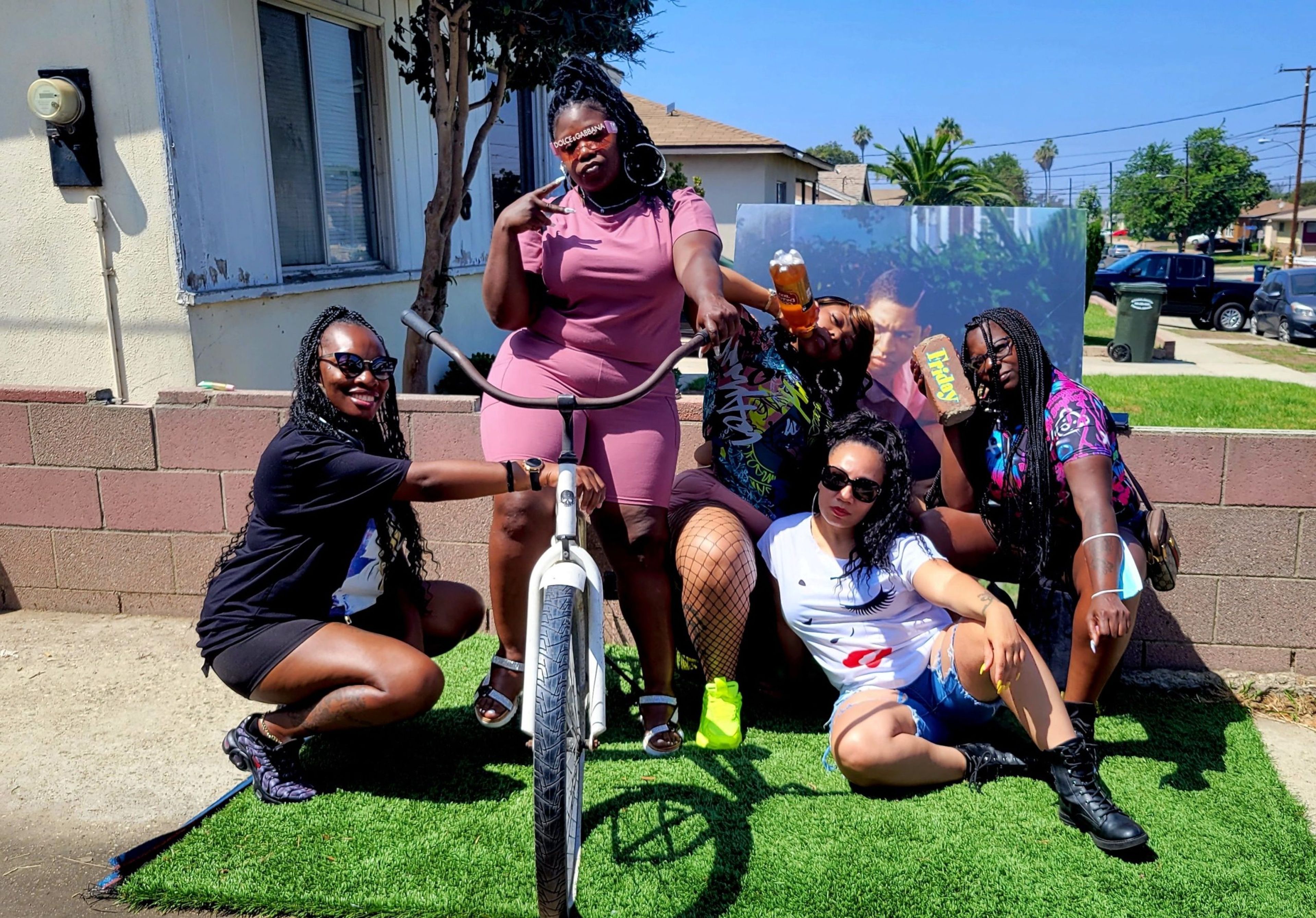 Group of five friends posing with a bicycle and drinks in a sunny outdoor setting.
