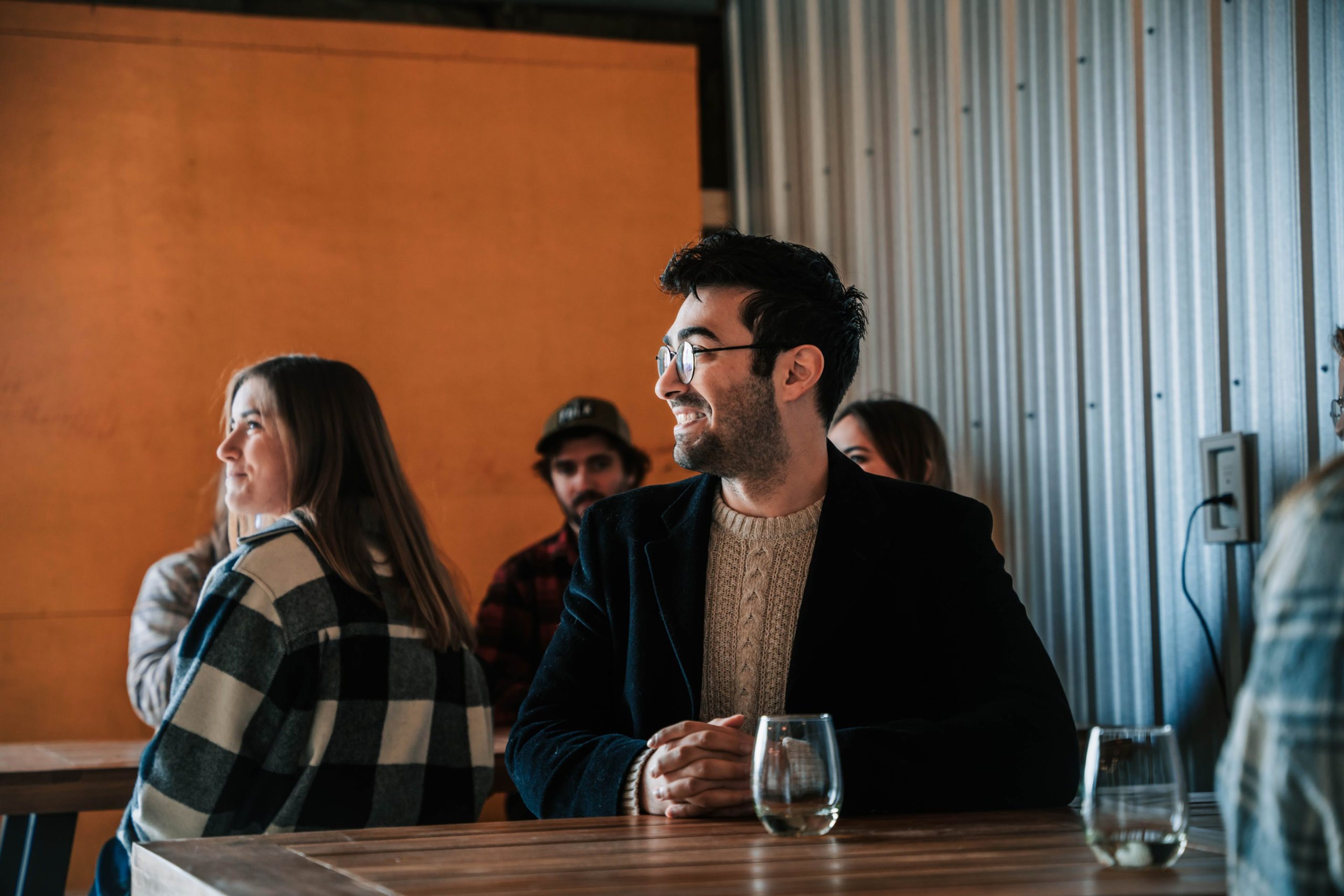 A group of people sitting at a table, one man smiling with glasses and a sweater.