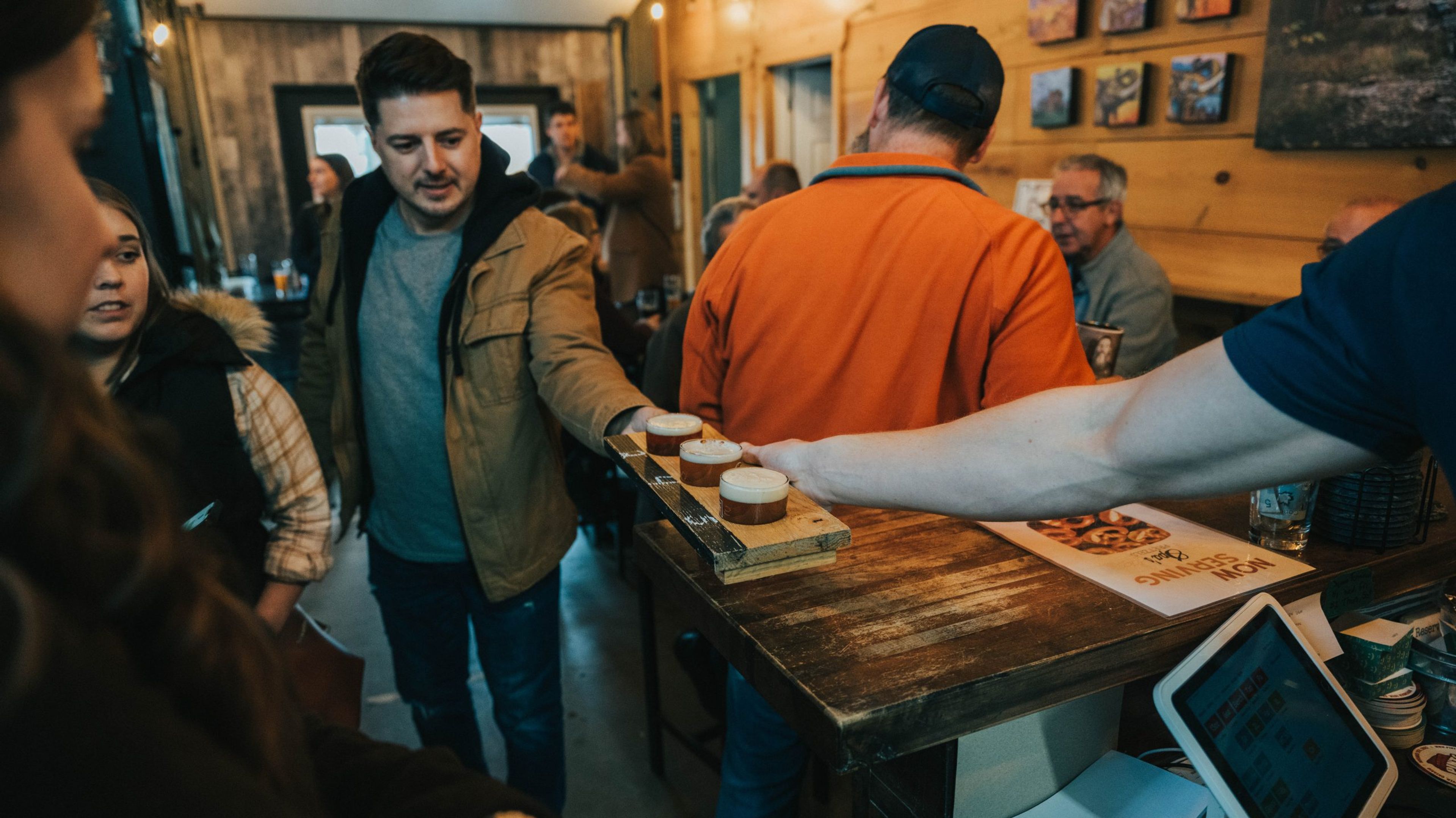 People enjoying drinks at a bar, with a server delivering a flight of beers.