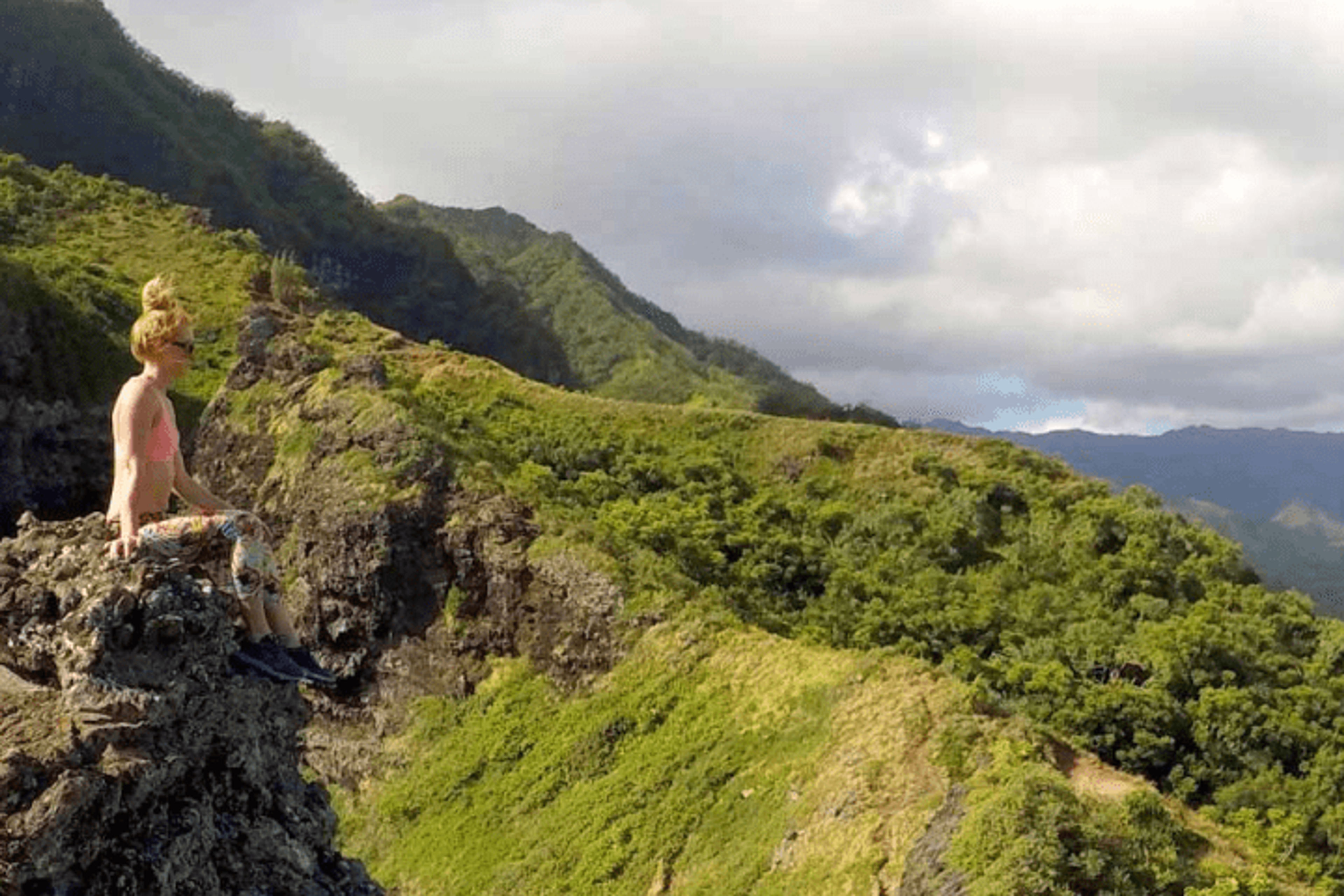 Person sitting on a rocky ledge overlooking green mountains and cloudy sky.
