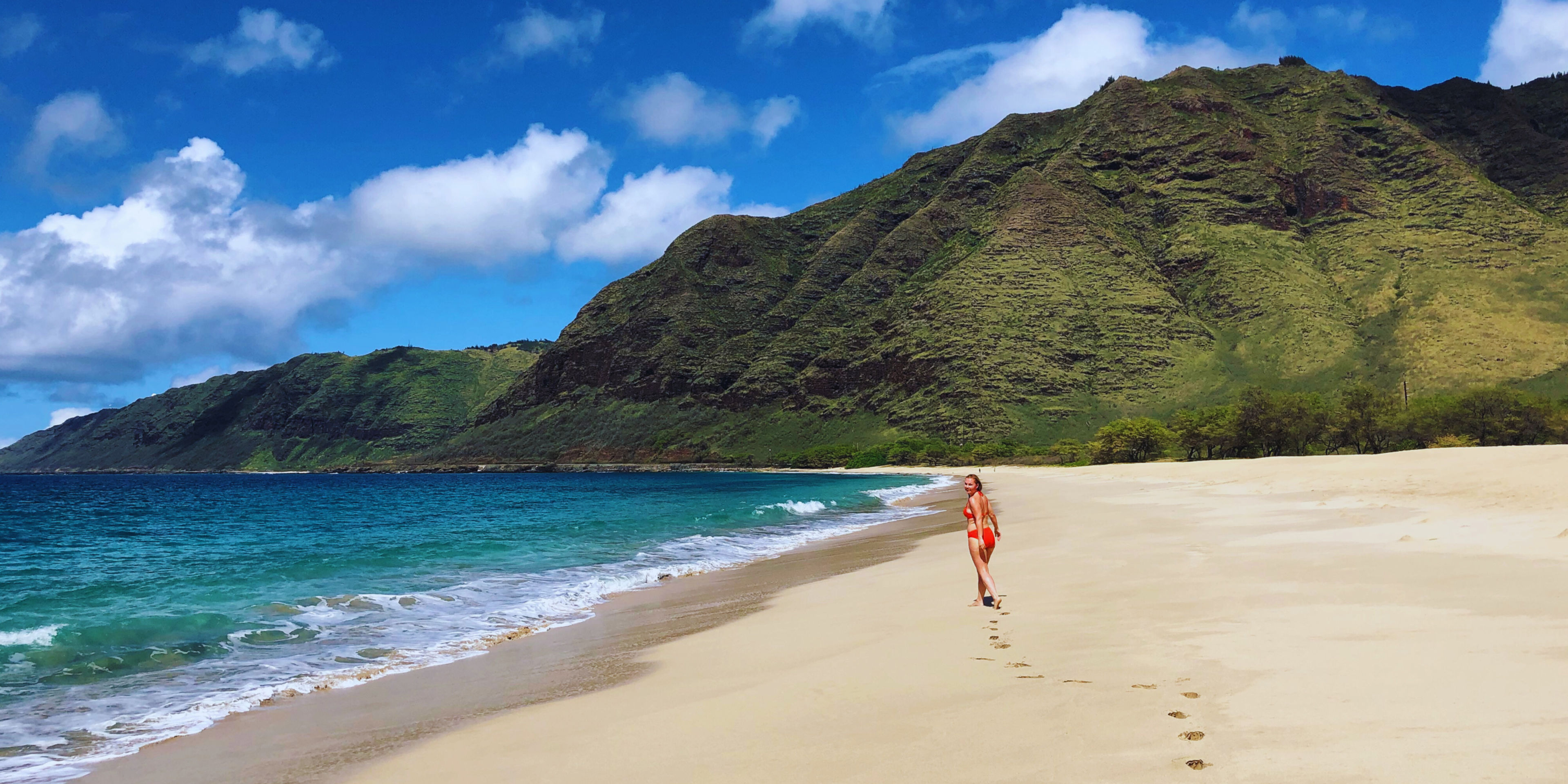 A person walking on a sandy beach with mountains and blue water in the background.