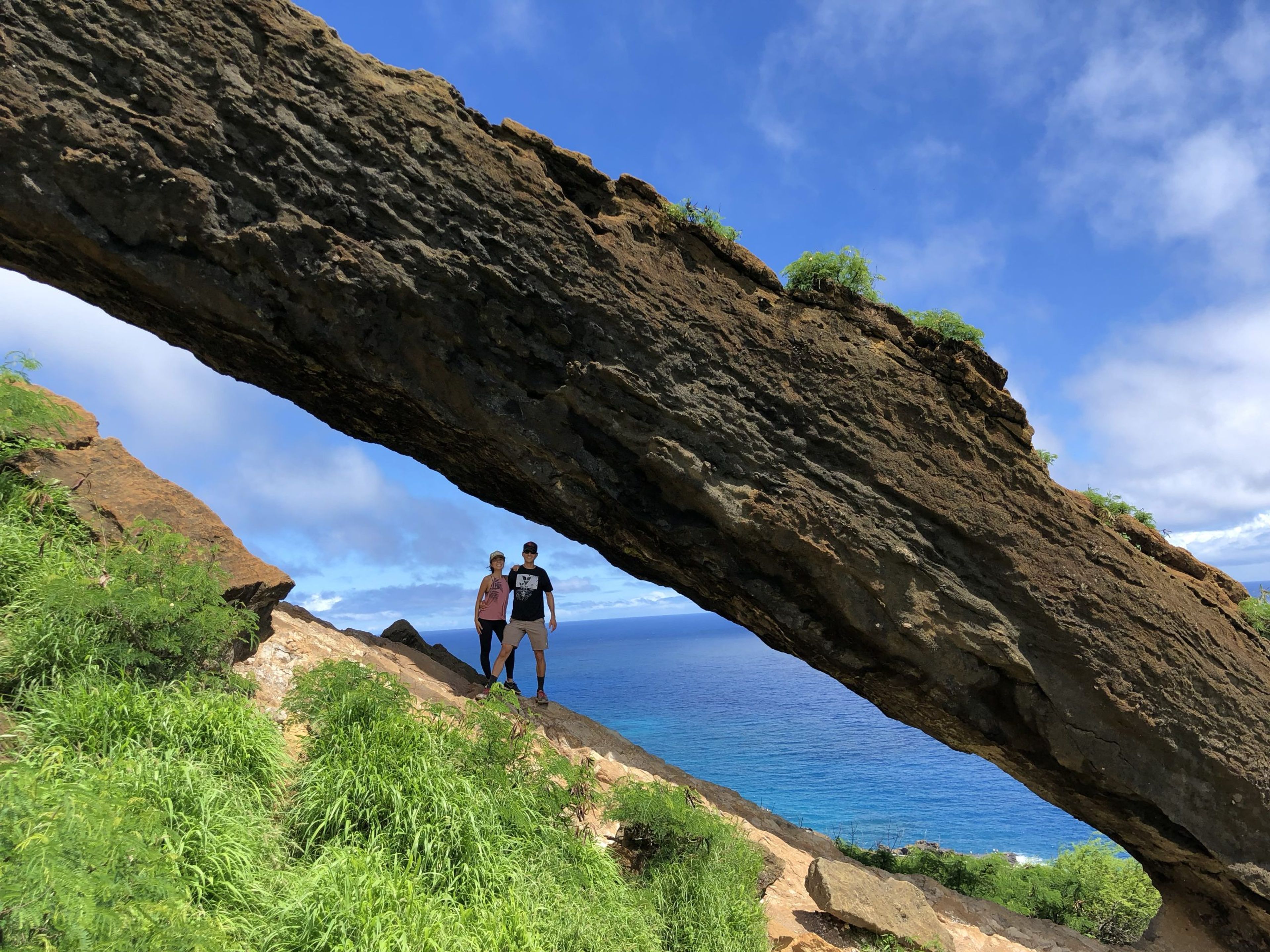 Two people standing under a rock arch with ocean view in the background.