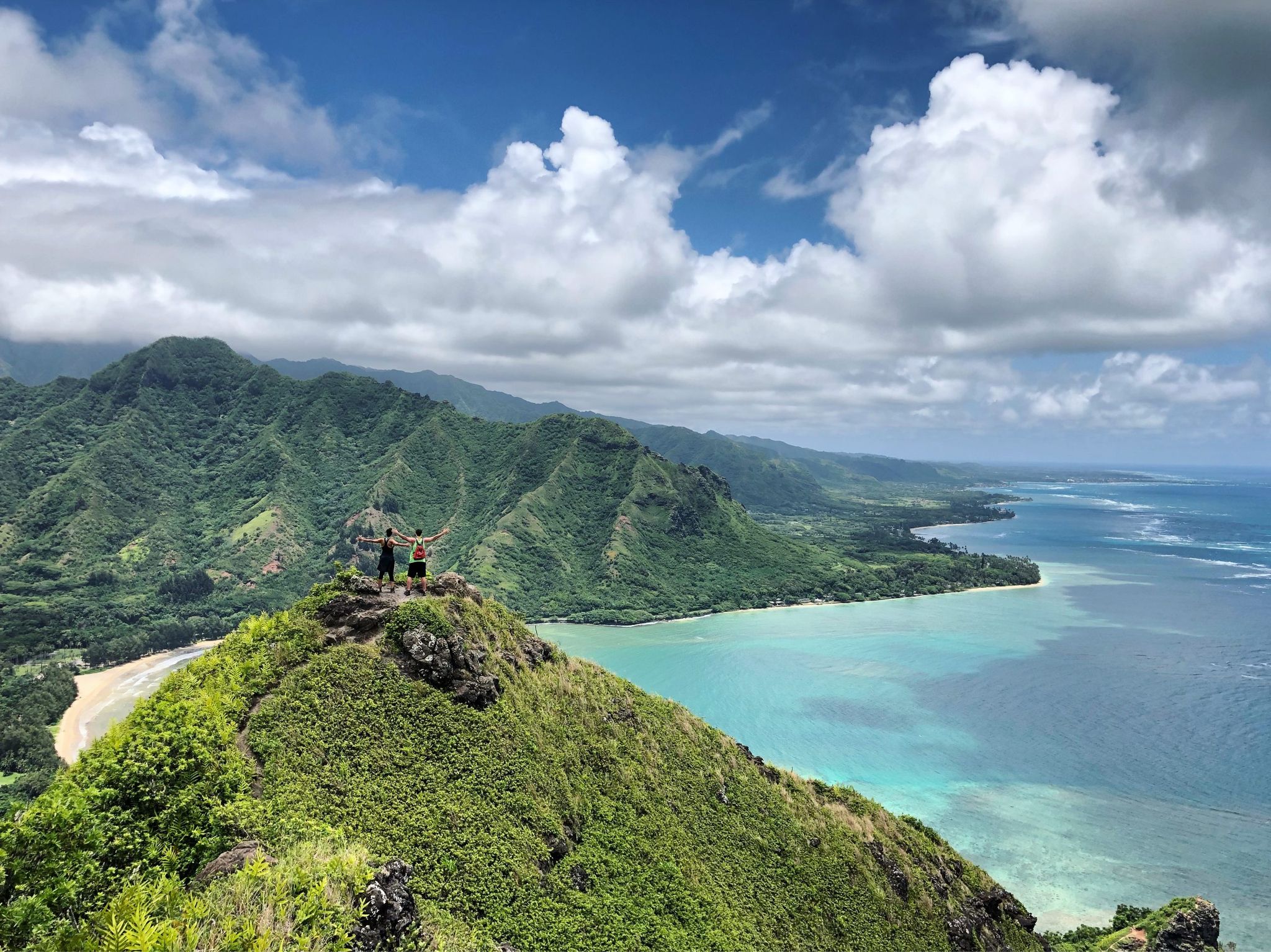 Two hikers on a mountain peak overlooking a lush green landscape and ocean.