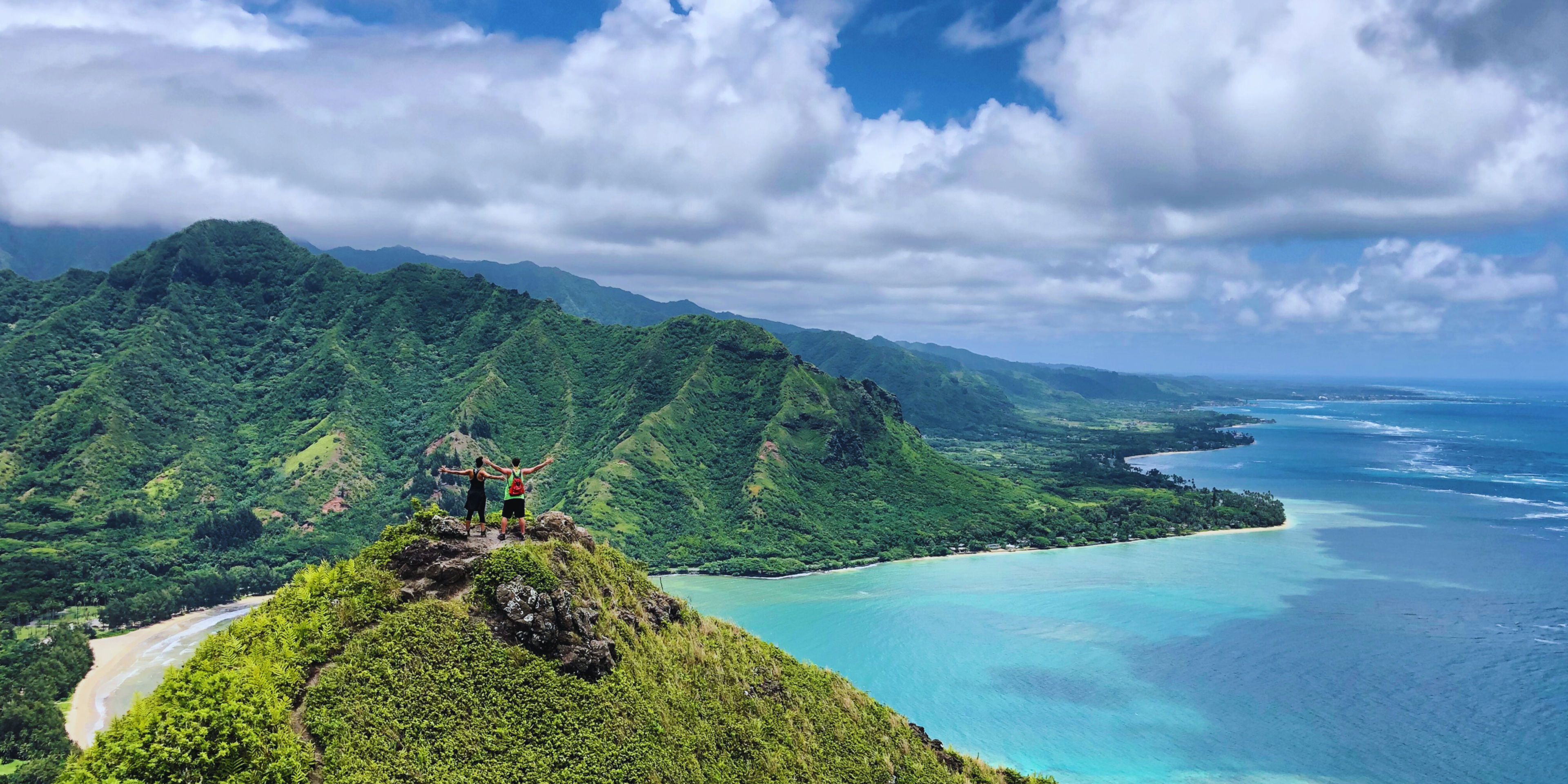 Two hikers celebrating on a mountain peak overlooking lush green hills and a blue ocean.