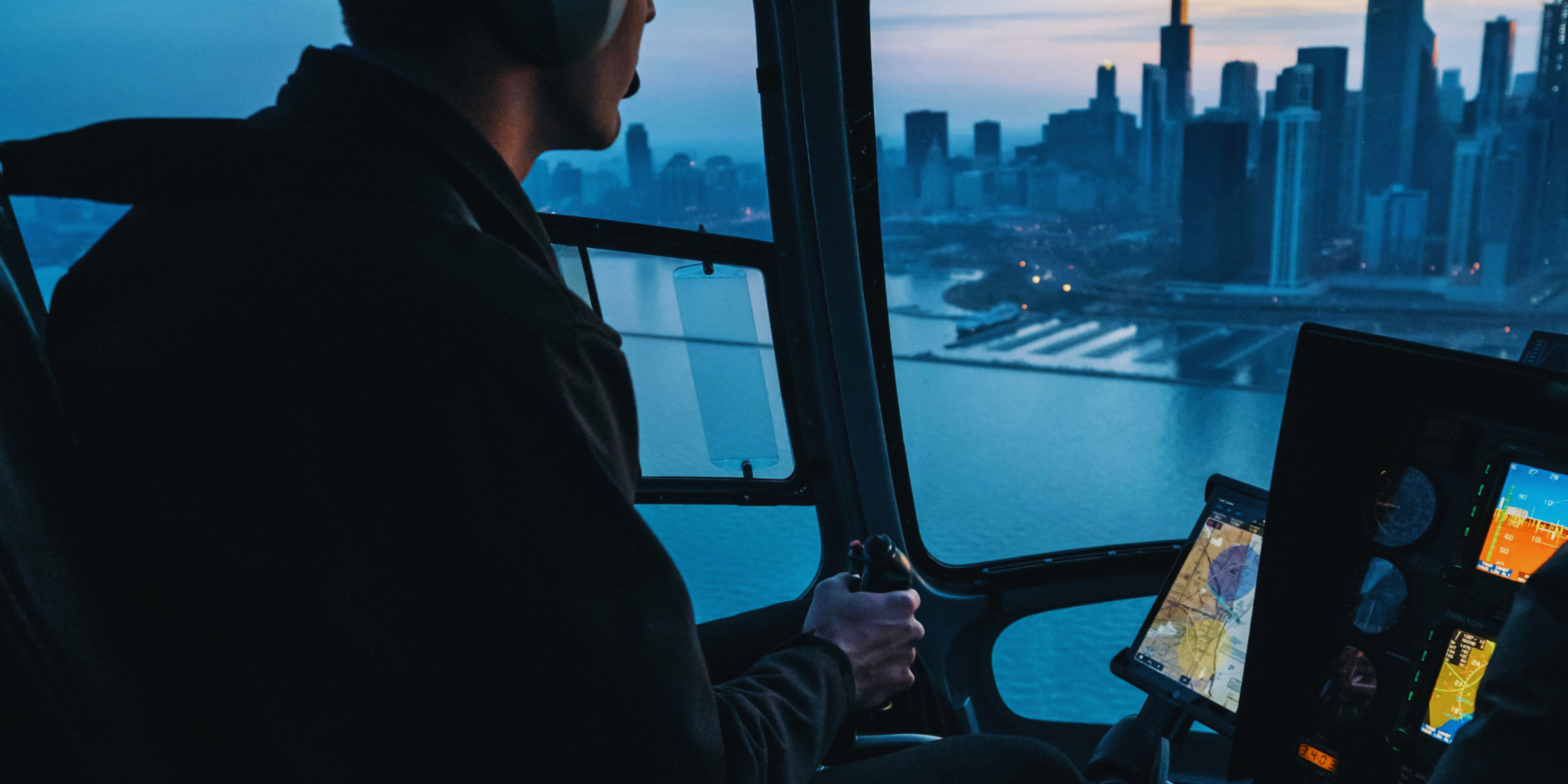 Pilot flying a helicopter over a city skyline at dusk.