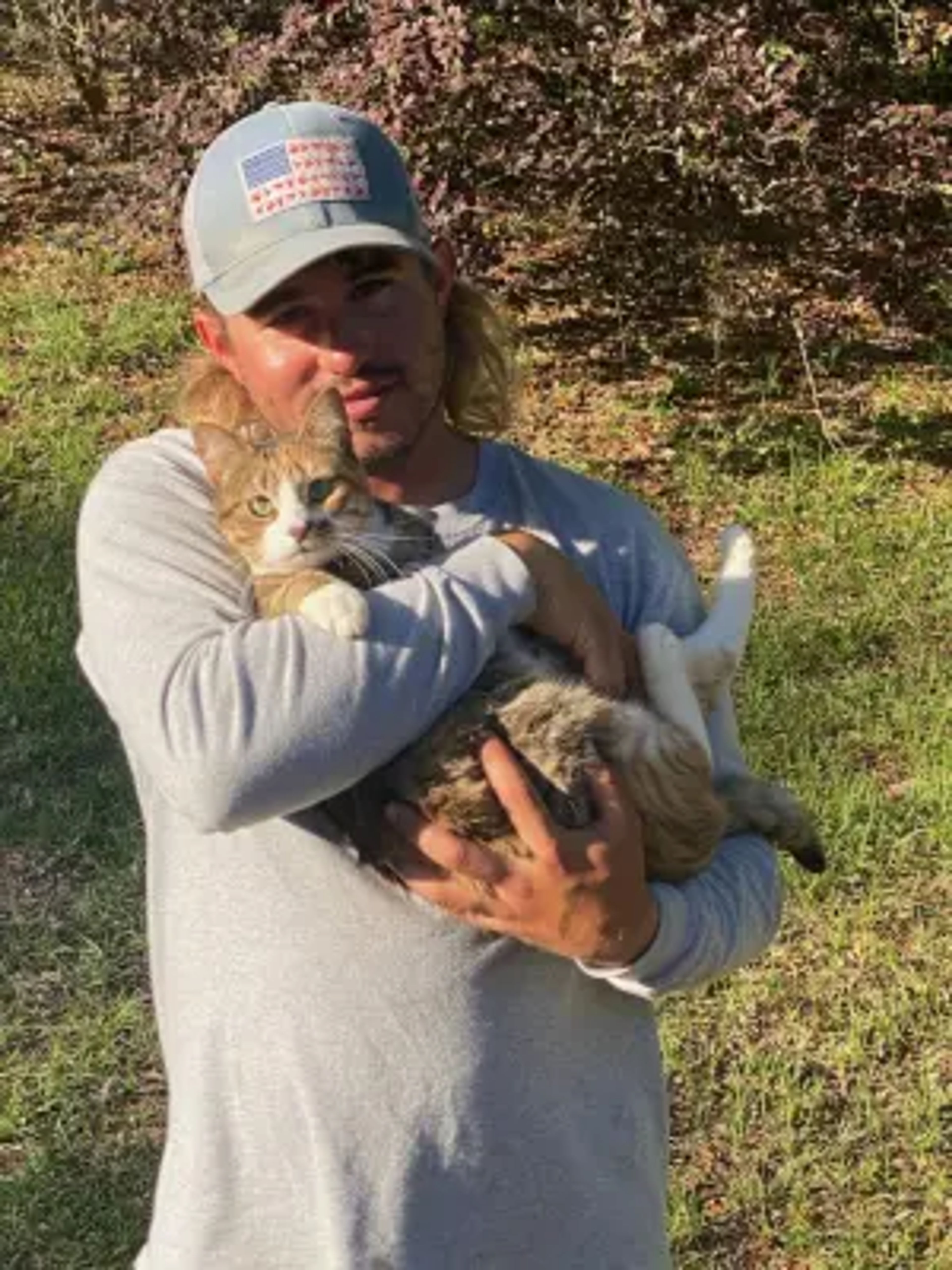 A person holding a cat outdoors with greenery in the background.