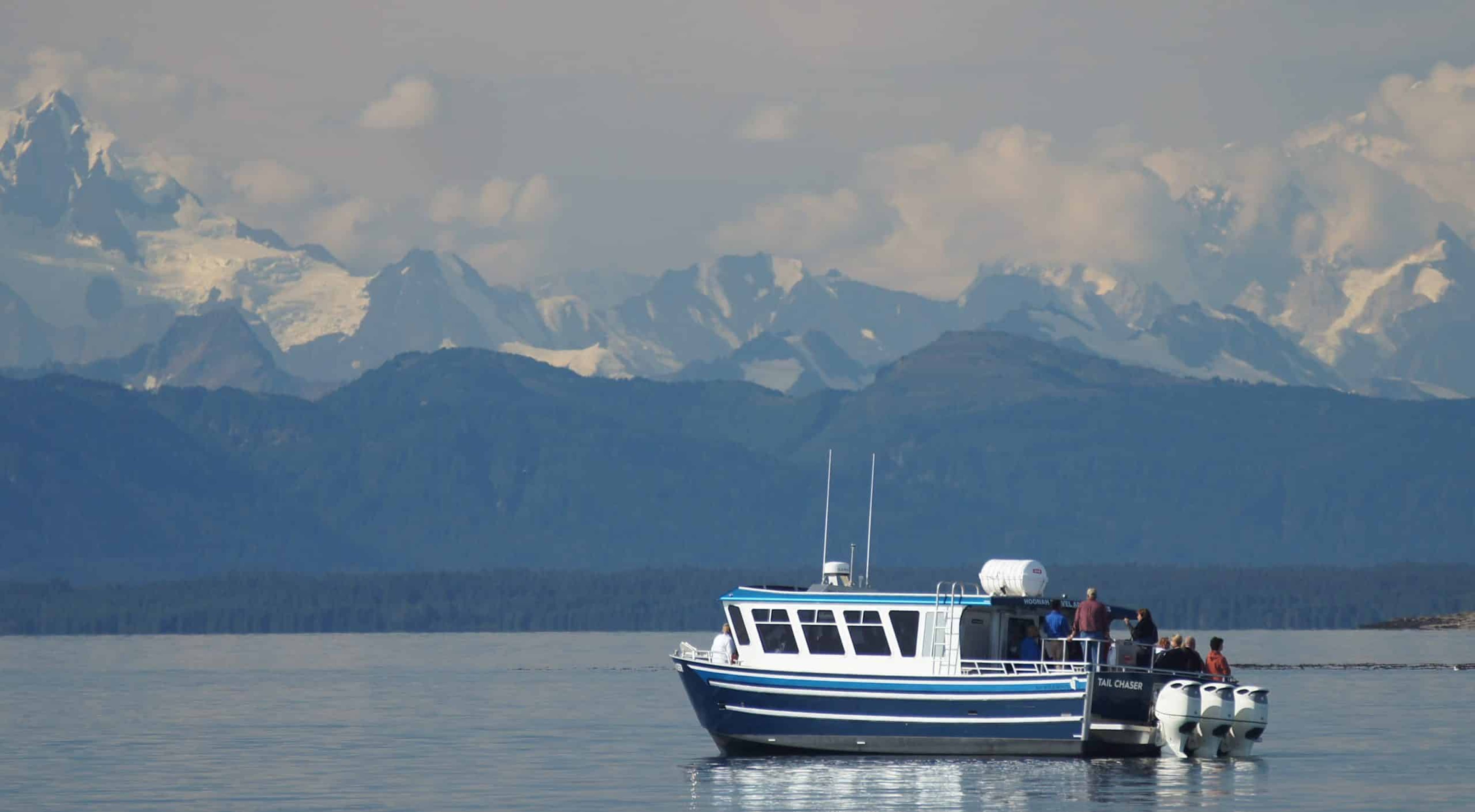 A blue boat with passengers on calm waters, surrounded by mountains and clouds.
