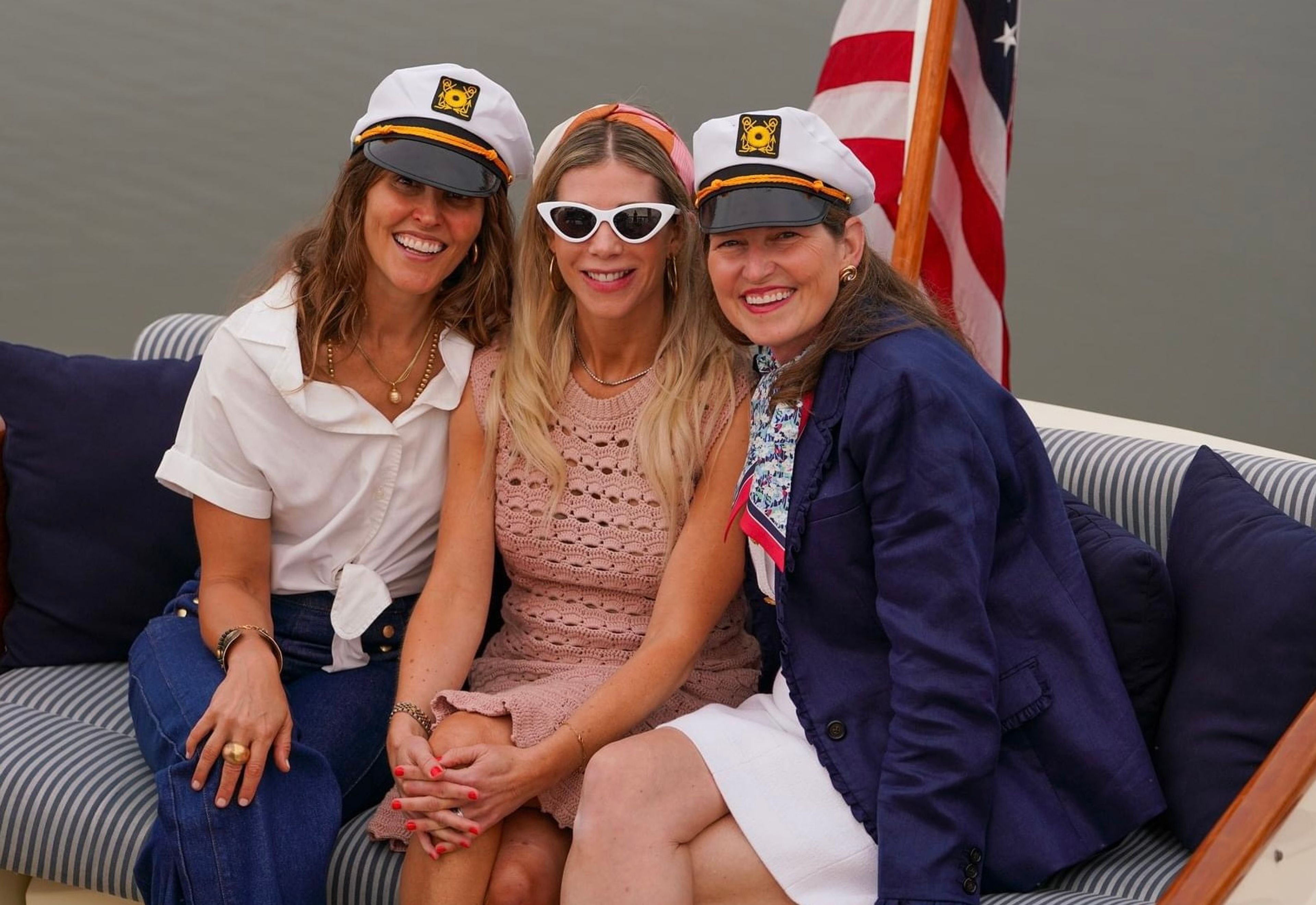 Three women in captain hats sitting together on a boat with an American flag in the background.