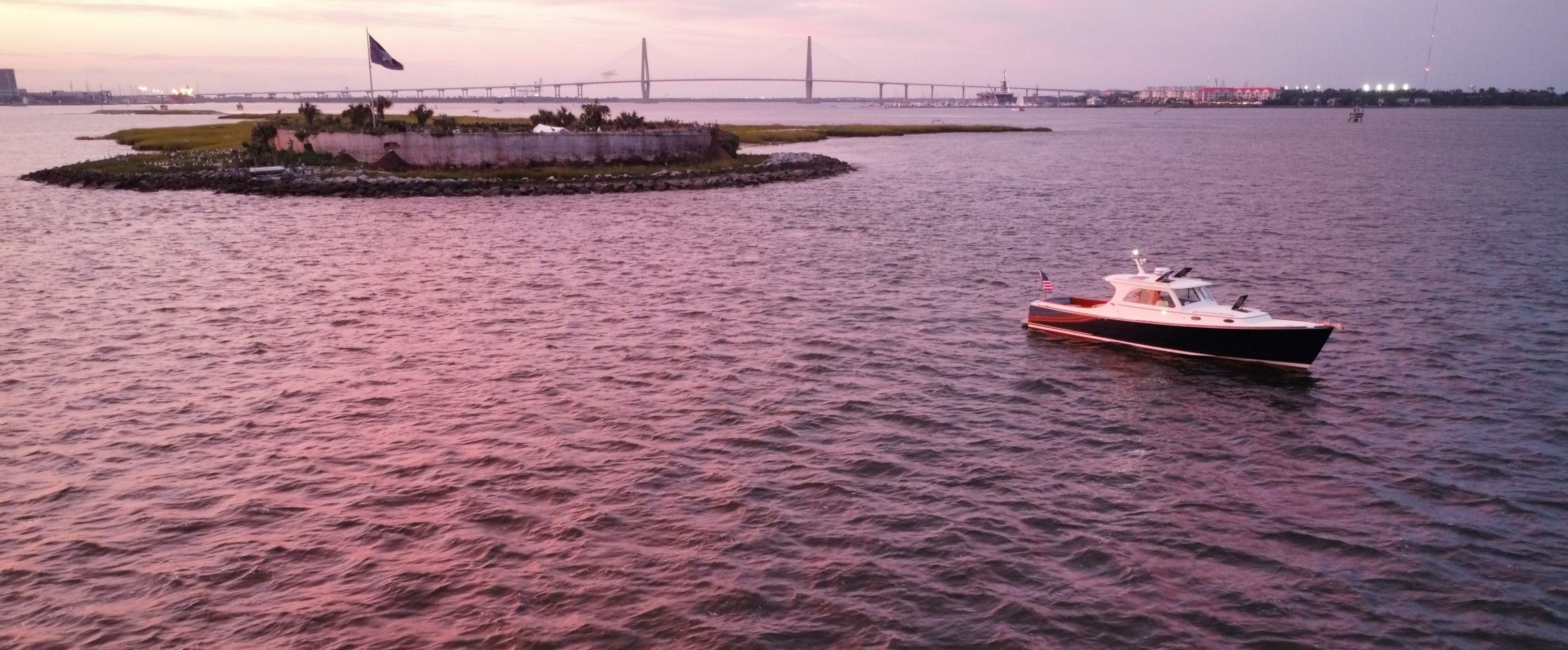 A boat on water with a bridge and island in the background during sunset.