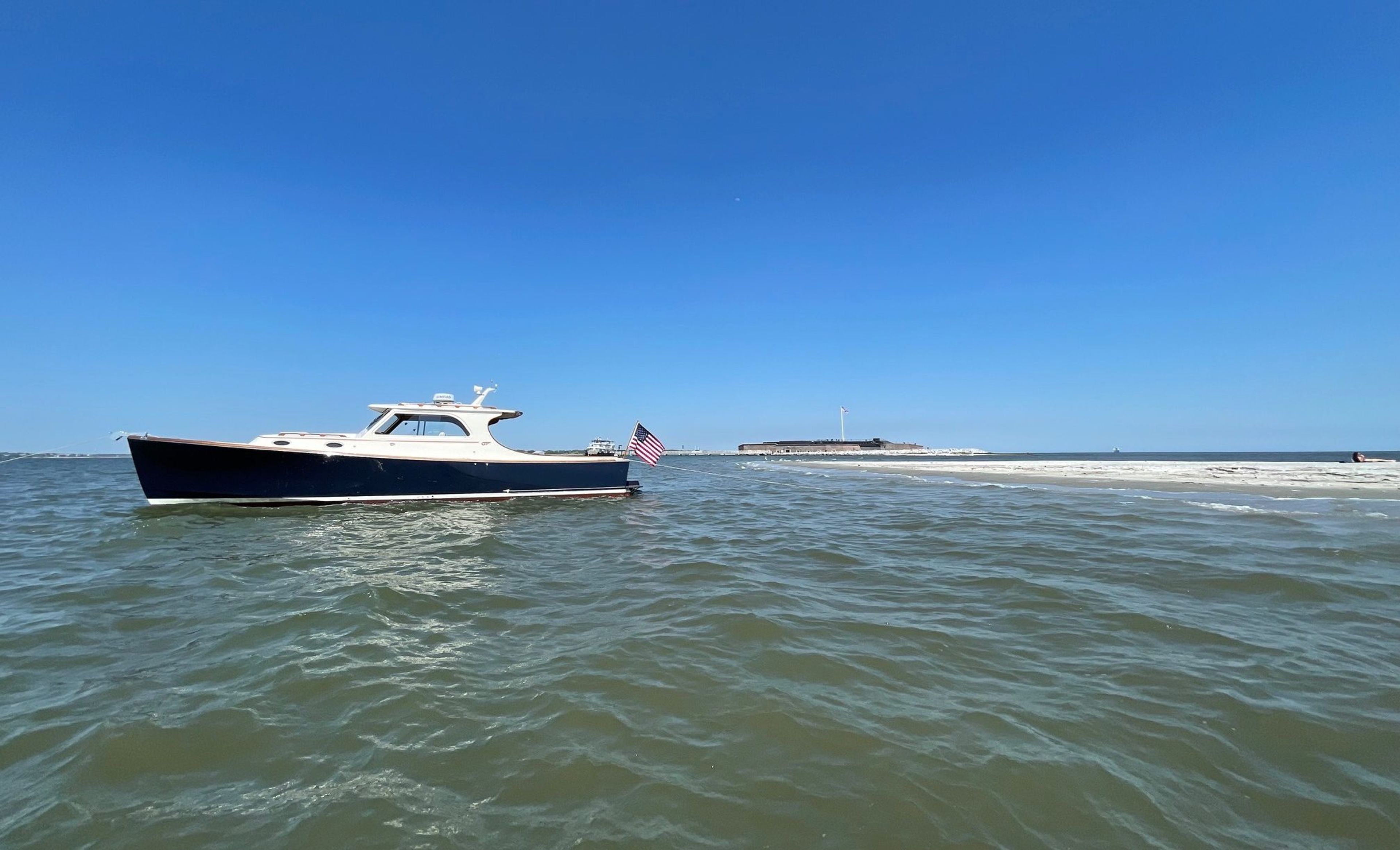 A boat with an American flag near a sandy shore and a fort under a clear blue sky.