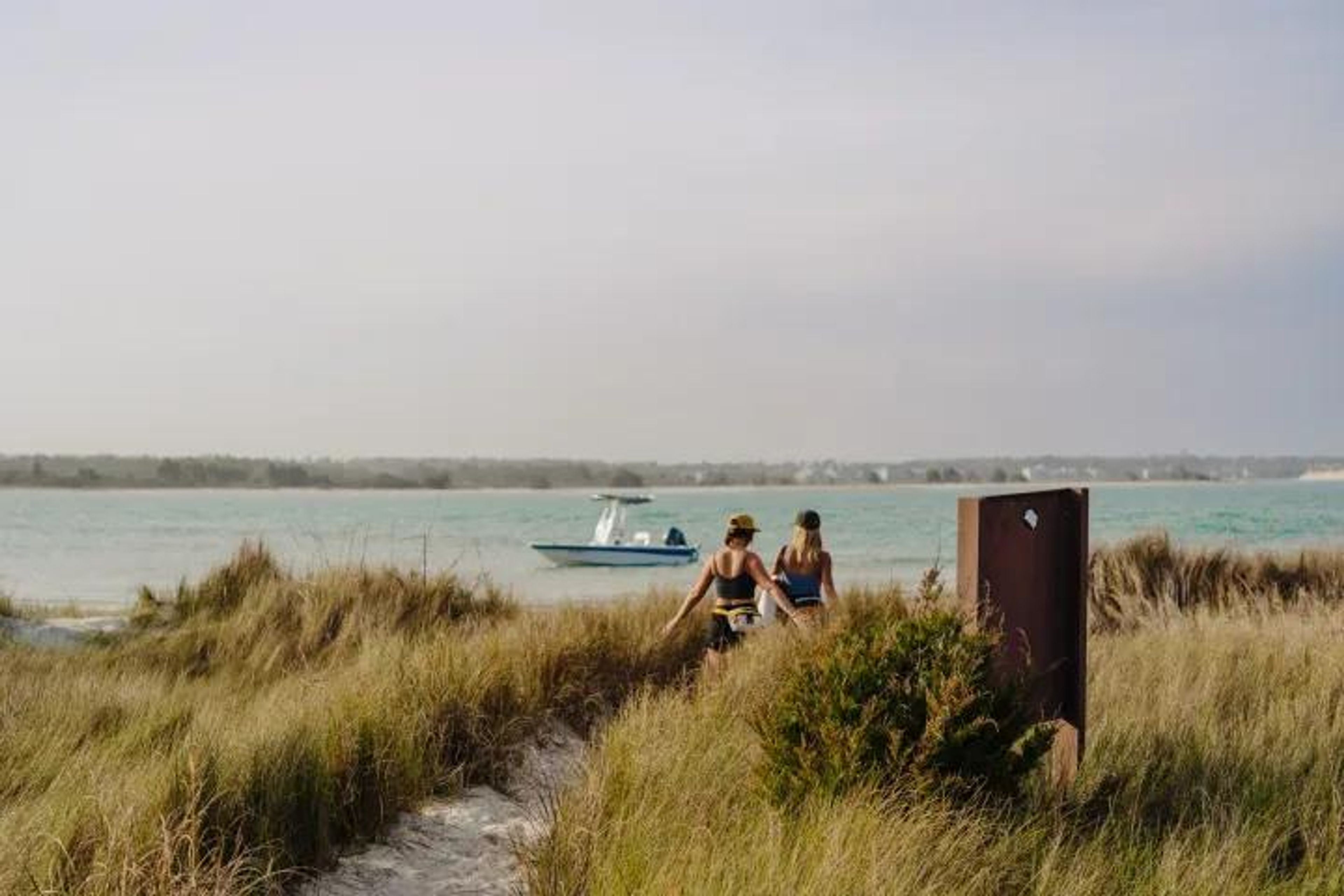 Two women walking on a sandy path towards the beach with a boat in the water.