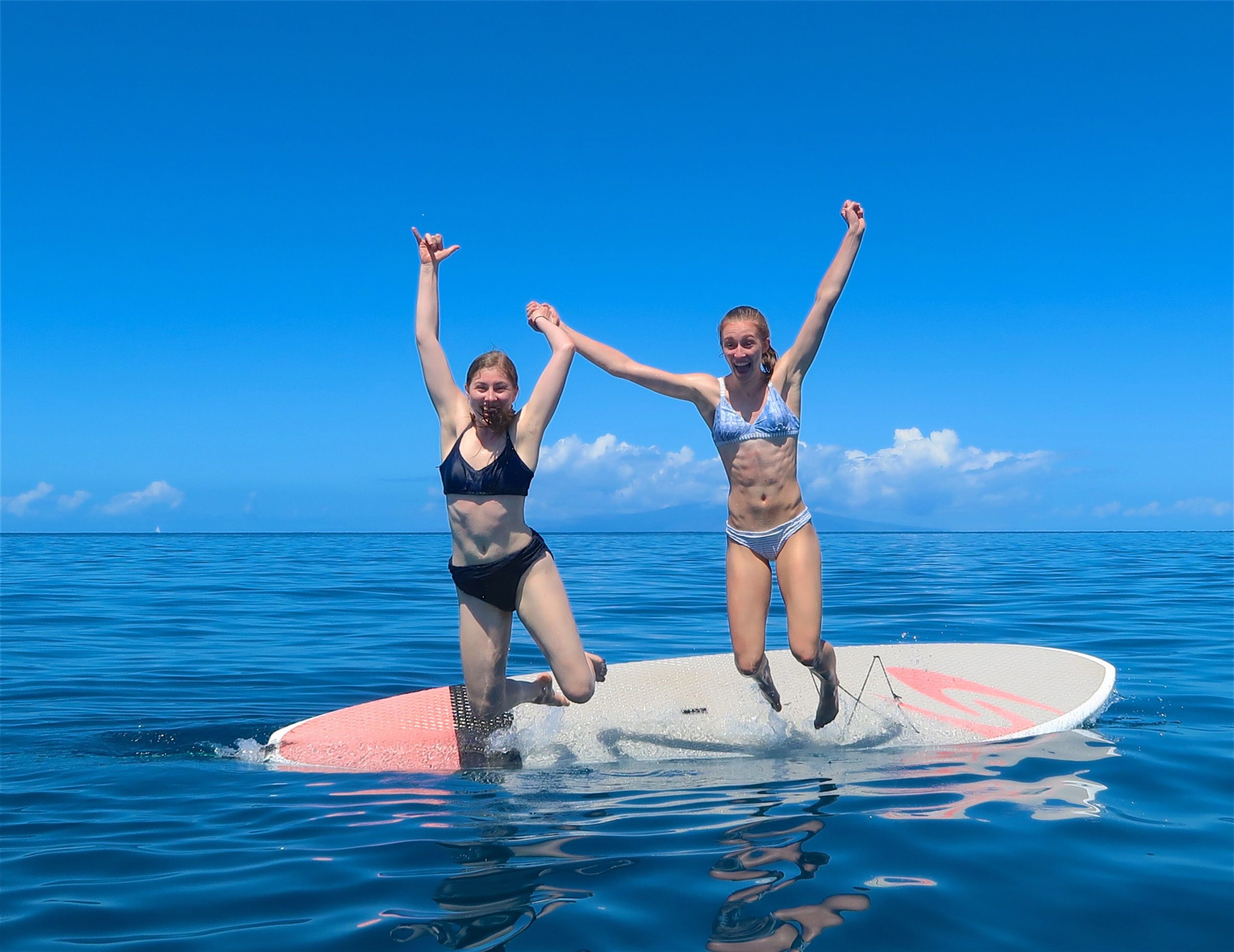 Two women jumping off a paddleboard in clear blue water under a sunny sky.