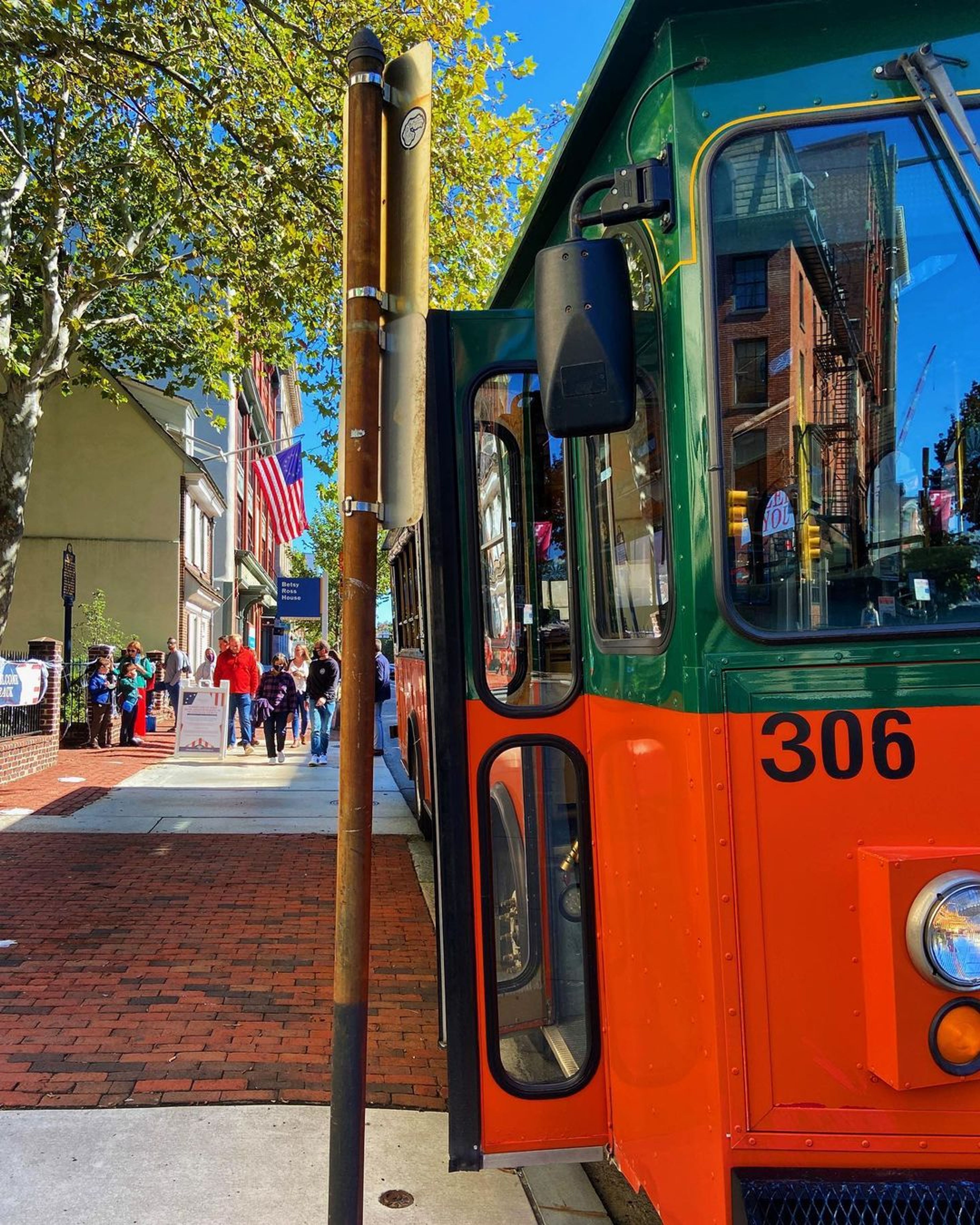 A colorful trolley bus parked near a busy street with people walking and trees in the background.