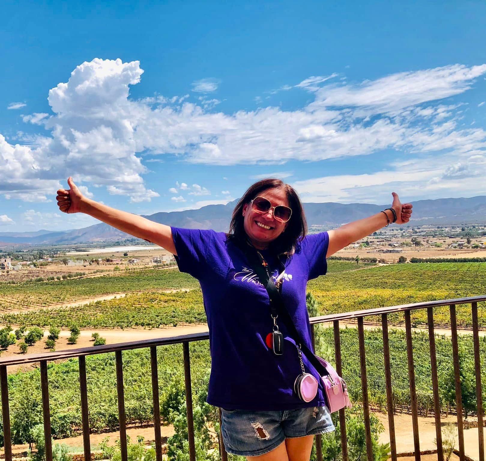 Person with arms raised in a vineyard with mountains and blue sky in the background.
