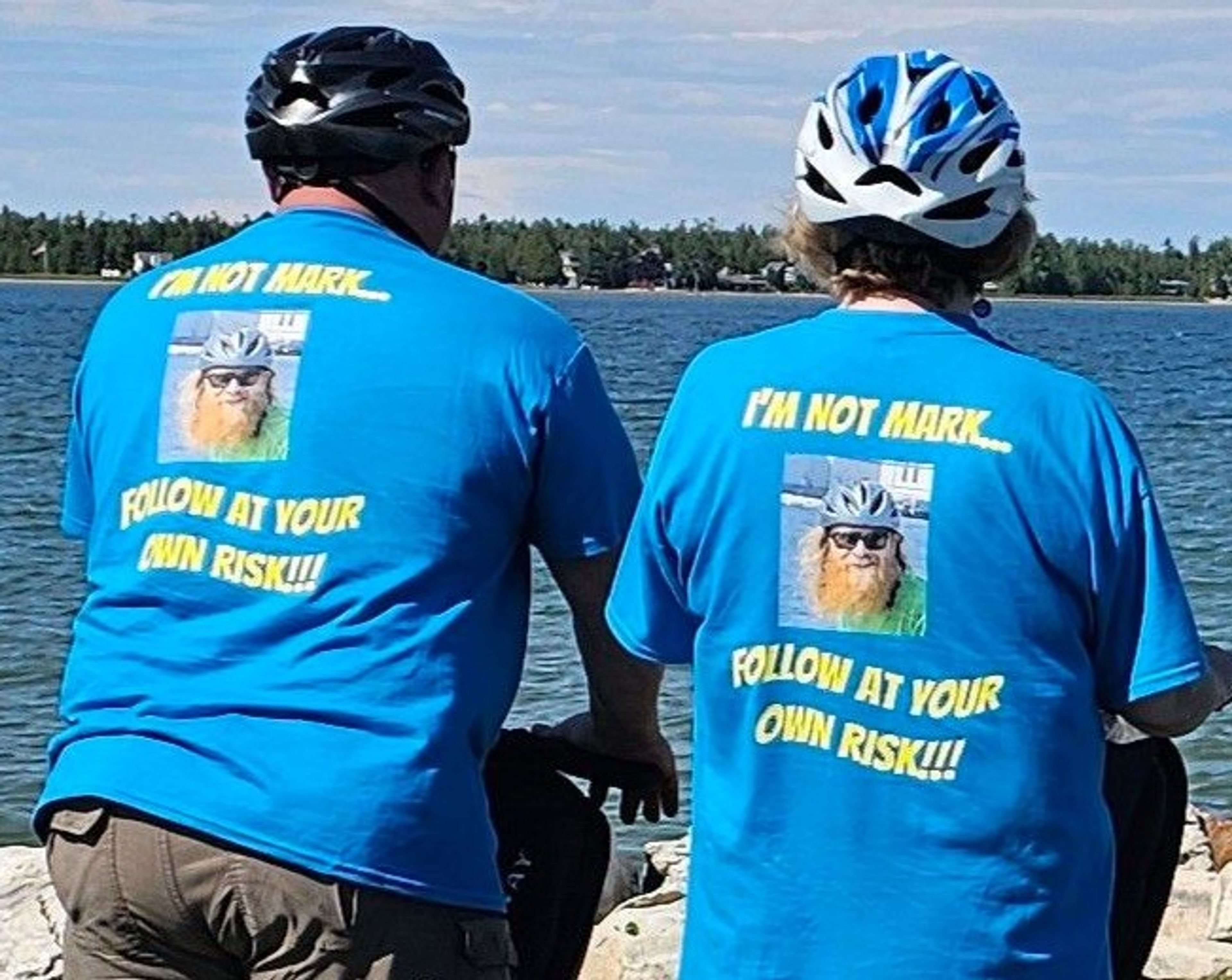 Two cyclists wearing matching blue shirts with humorous text by a lake.