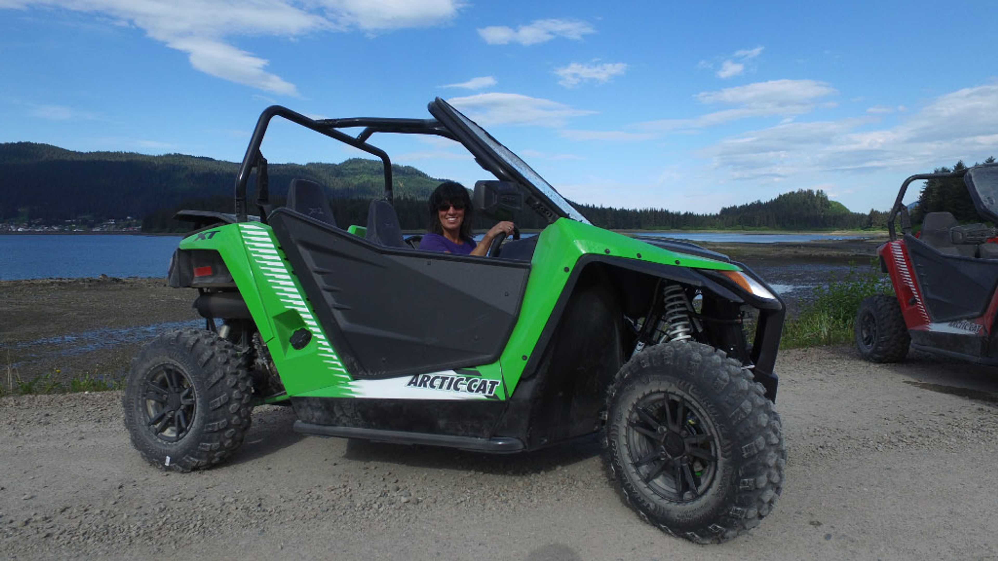 A person sitting in a green Arctic Cat off-road vehicle by a lake with mountains in the background.