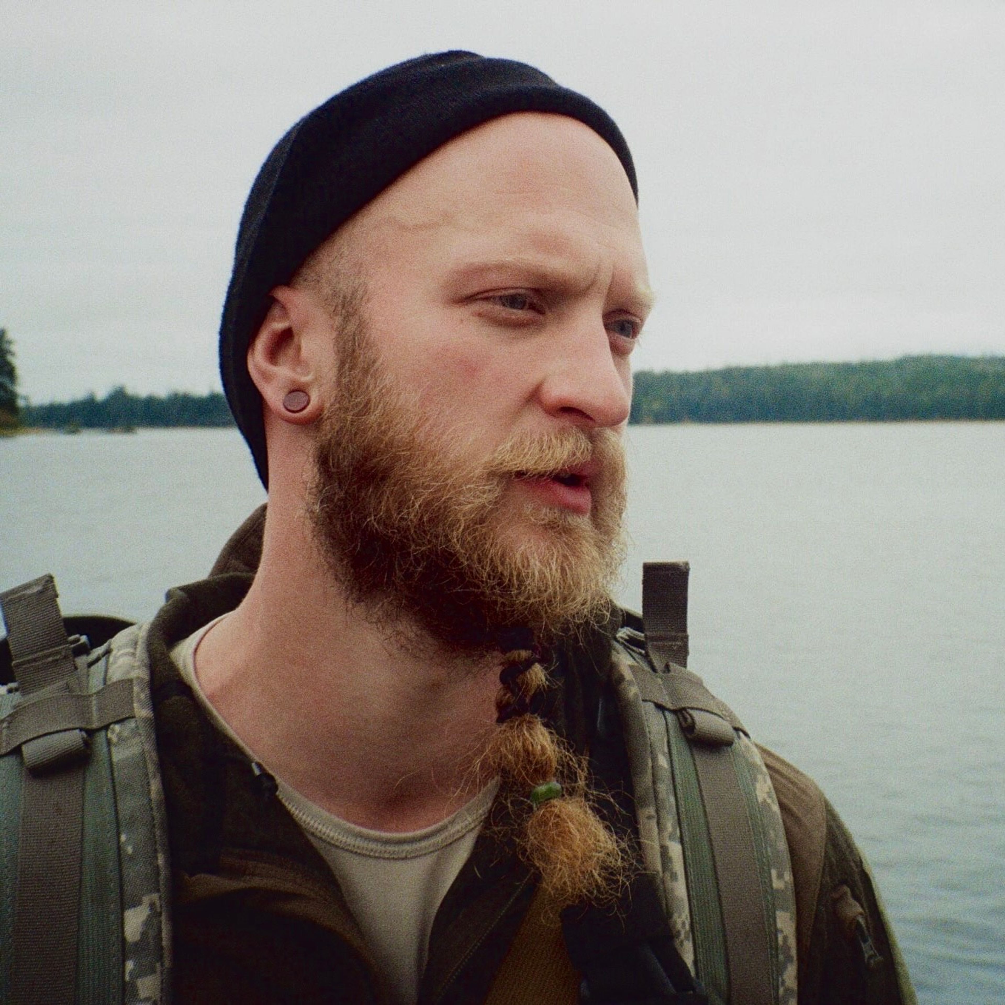 Man with a beard and beanie standing by a lake, wearing a backpack.