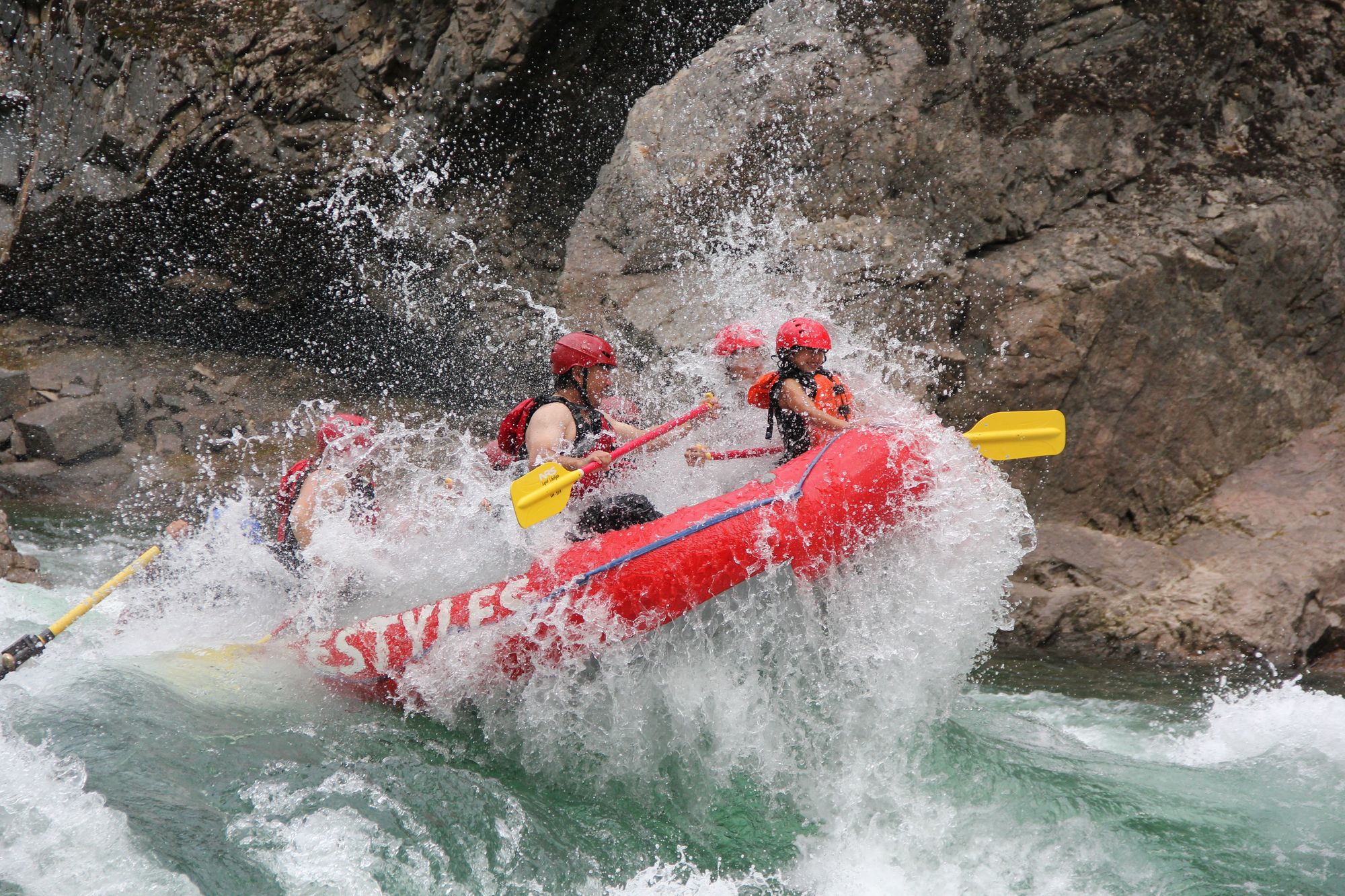 Family on the whitewater trip splashing over a rolling wave making huge water splash back. You kids at the helm bracing the wave