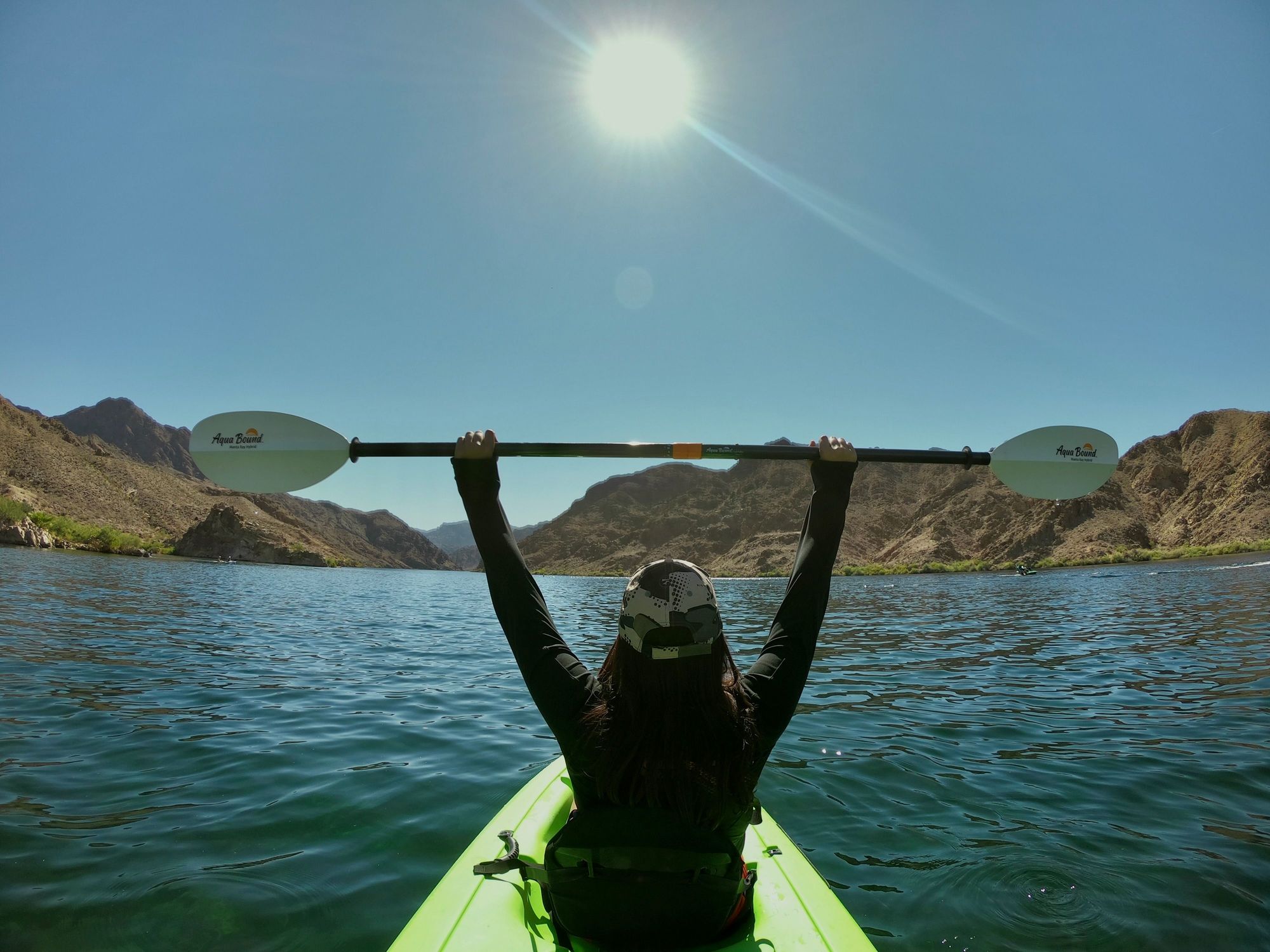 A solo kayaker paddles through calm turquoise waters surrounded by rugged desert canyon walls under a clear blue sky.