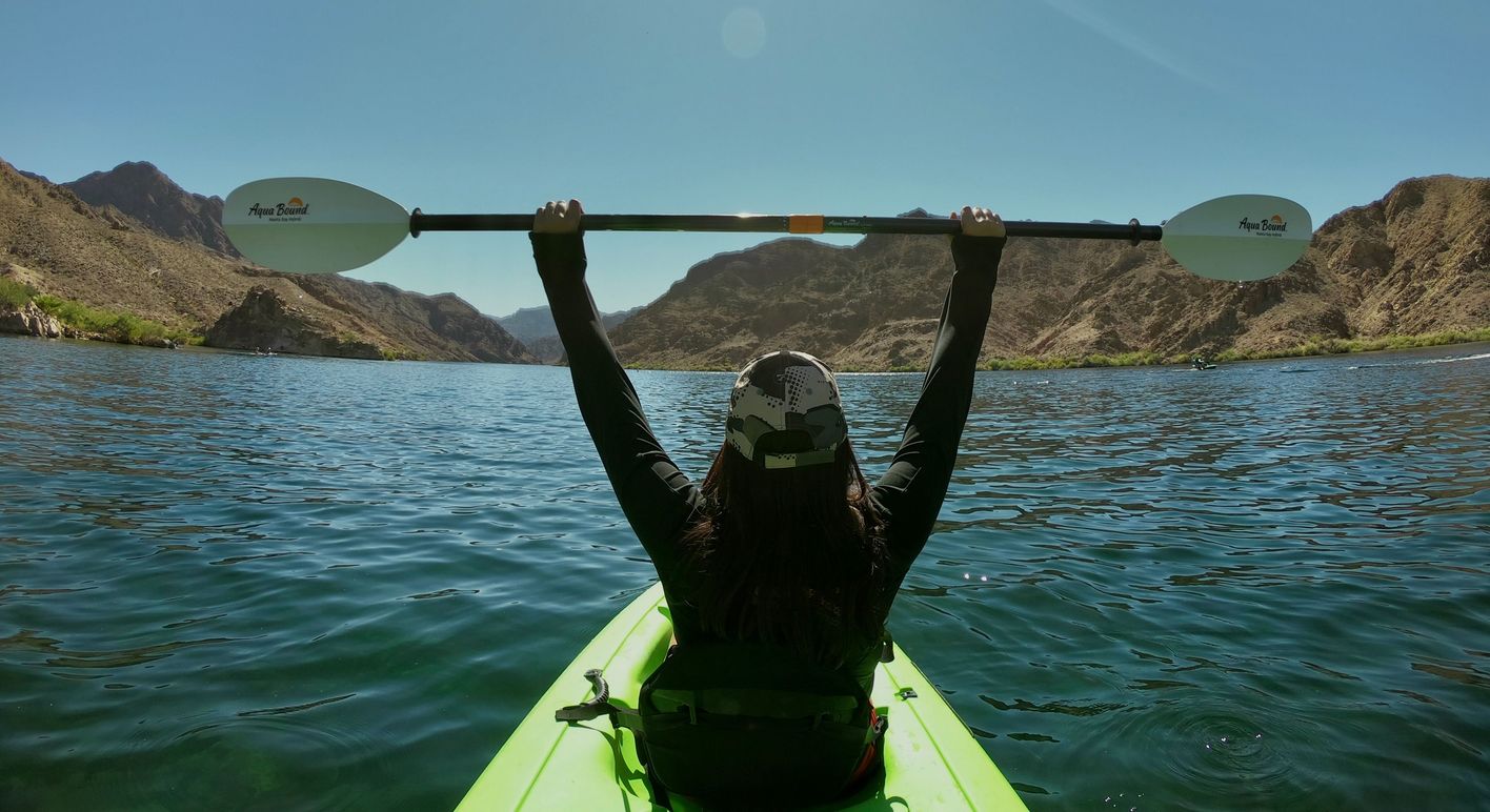 A solo kayaker paddles through calm turquoise waters surrounded by rugged desert canyon walls under a clear blue sky.