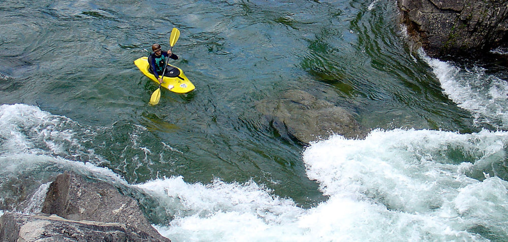 Someone in a yellow kayak navigating the rapids. The water is deep green with the rocks surrounding the sides of the boater. 