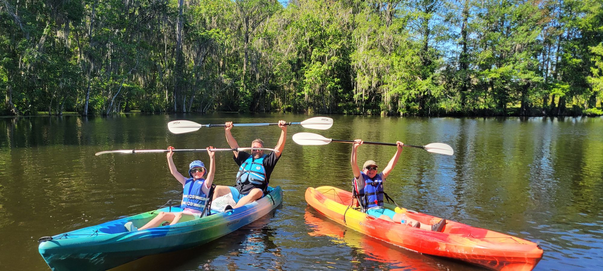 Three kayakers celebrate on the Dora Canal, raising their paddles in excitement as they float on calm waters surrounded by lush cypress trees.