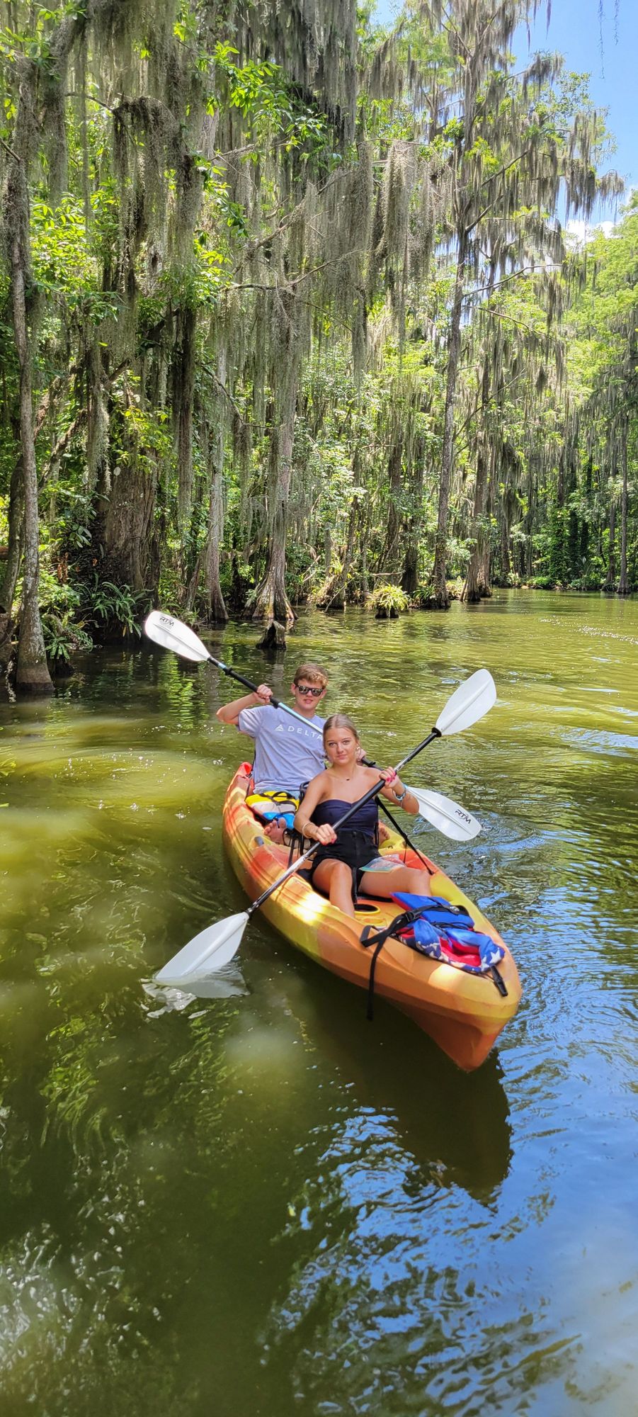 Two kayakers paddle through the scenic Dora Canal, surrounded by towering cypress trees draped in Spanish moss. The calm waters and lush greenery make for a perfect outdoor adventure in Florida.