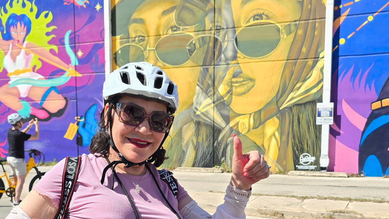 A smiling woman wearing a bike helmet and sunglasses poses in front of a vibrant mural in San Antonio&rsquo;s D.R.E.A.M. District during a Mural Ride Bike Tour.