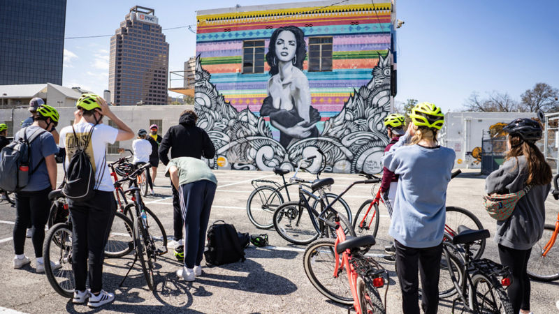 High school students on a Mural Ride bike tour stop to observe a vibrant Los Otros mural in downtown San Antonio, featuring a bold black-and-white portrait framed by colorful Latin-inspired stencils and signature filigree linework.