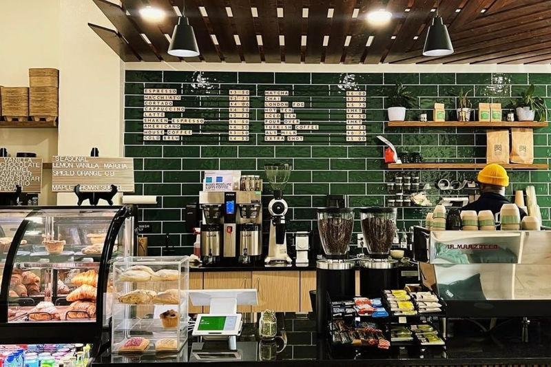 A stylish coffee shop with a dark green tiled wall, featuring a minimalist menu board displaying espresso drinks. A barista in a yellow beanie works behind the counter, which is lined with fresh pastries, coffee grinders, and an espresso machine.