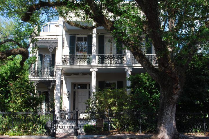 Large white mansion with Greek revival columns and intricate cast ironwork. Large Southern live oak tree in front.