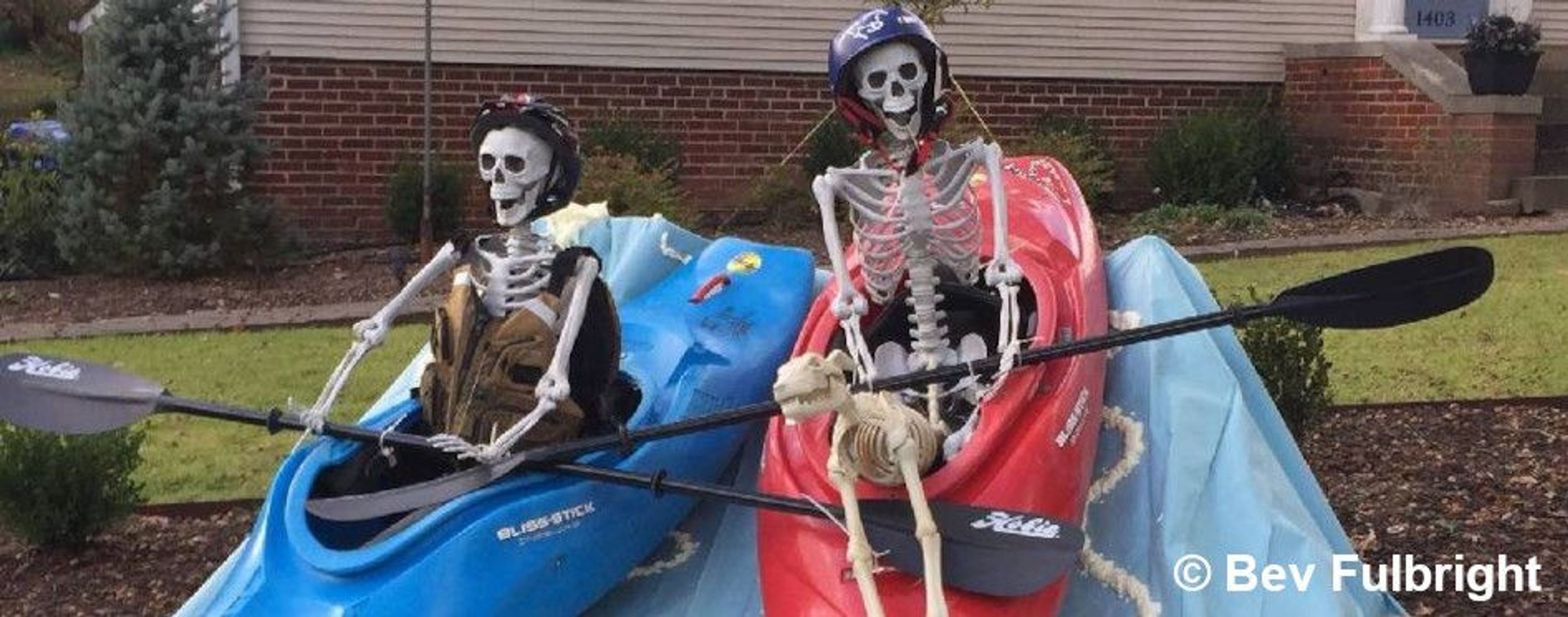 Two skeleton decorations dressed in kayaking gear and helmets sitting in colorful kayaks for a spooky Halloween kayaking display