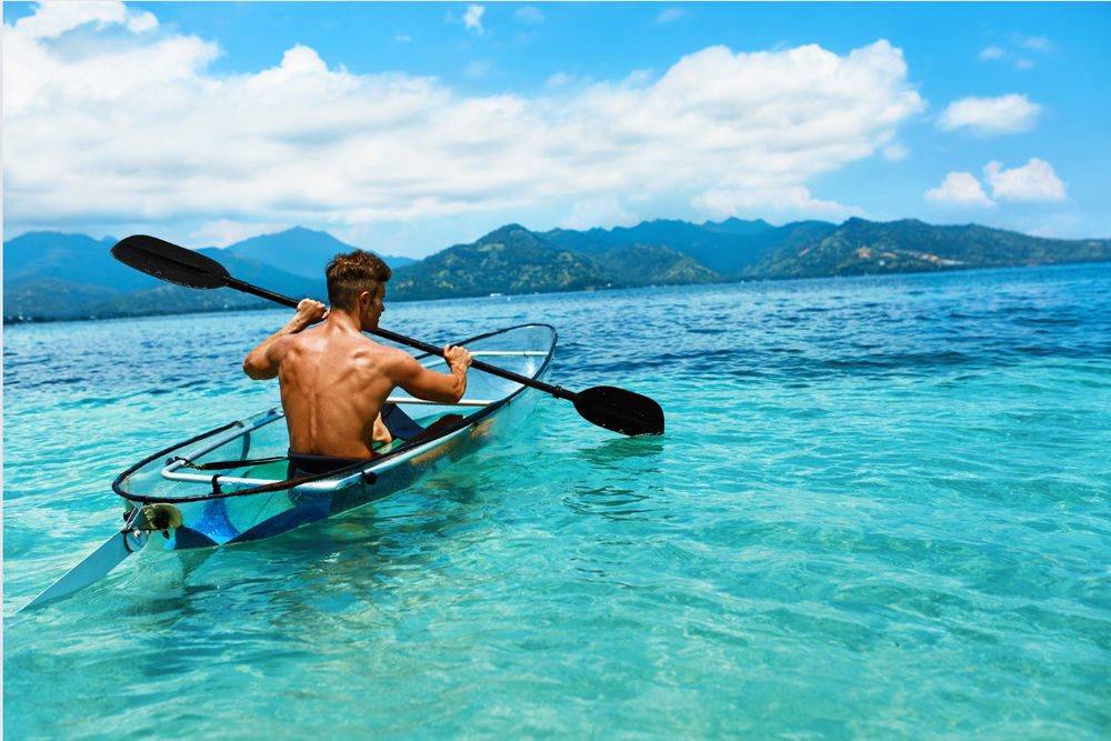 Male person paddling in a clear kayak in crystal clear waters and mountains in the background.