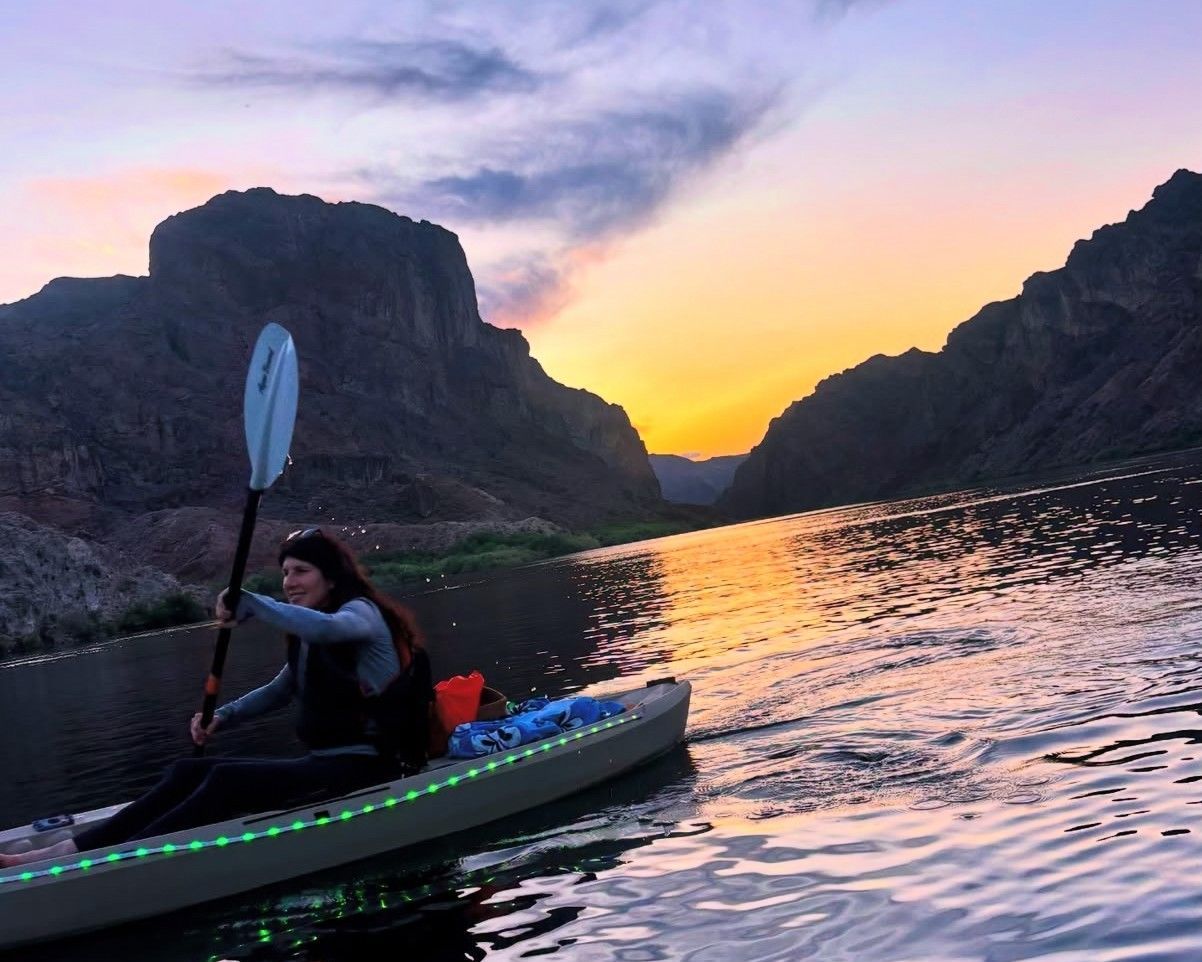 A kayaker paddles peacefully on the Colorado River at sunset with canyon walls glowing in golden light.