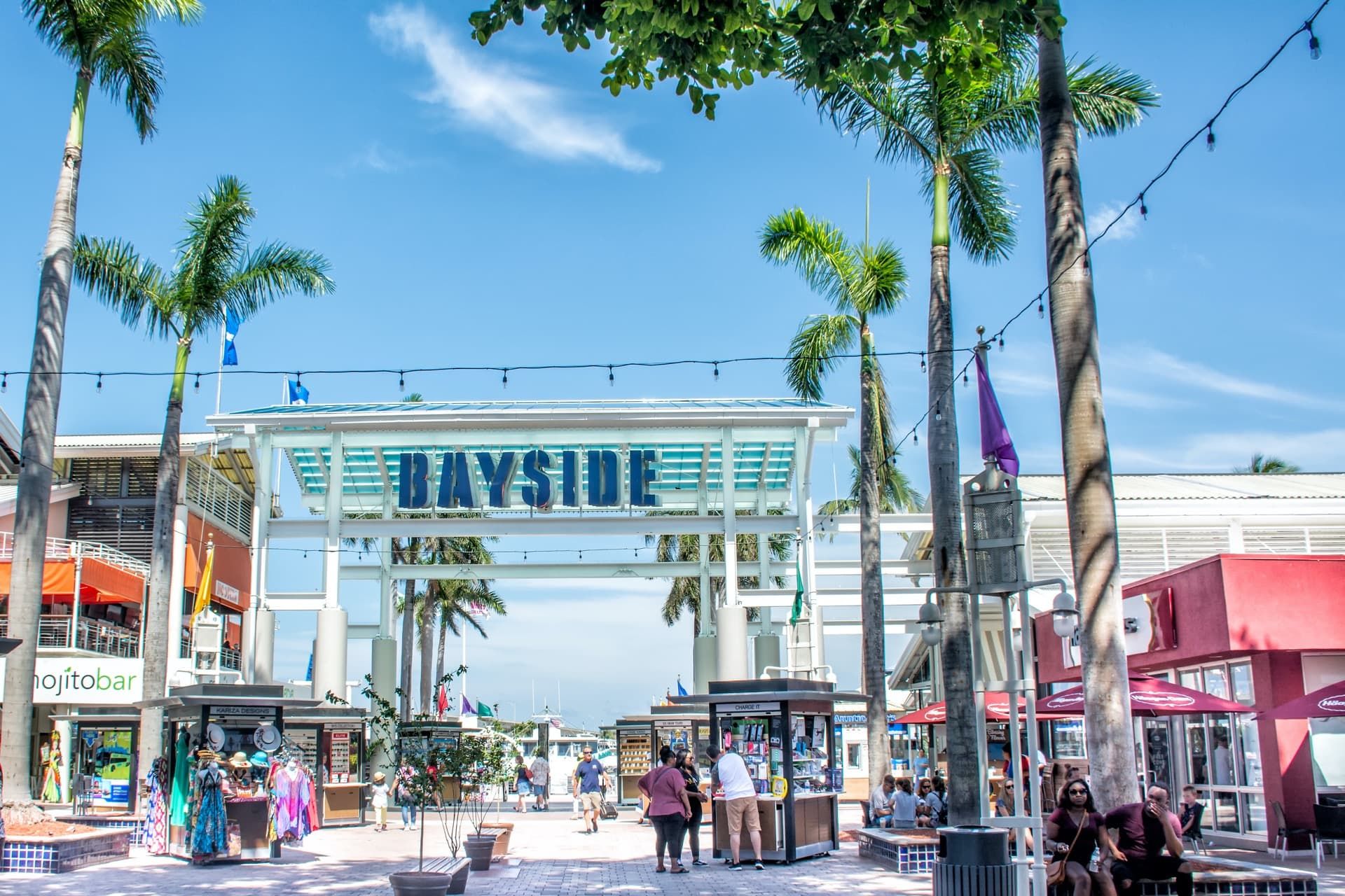 Bustling walkway at Bayside Marketplace with visitors, palm trees, and shops under a clear blue sky.