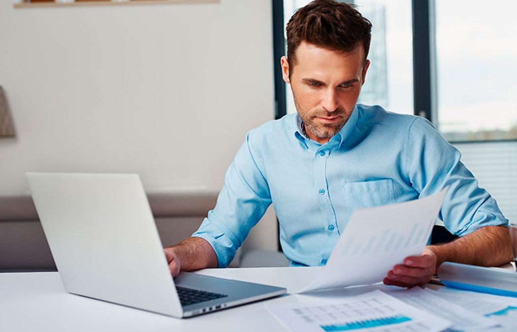 Foto de um homem com camisa social azul sentado lendo uma folha e com um notebook ao lado.