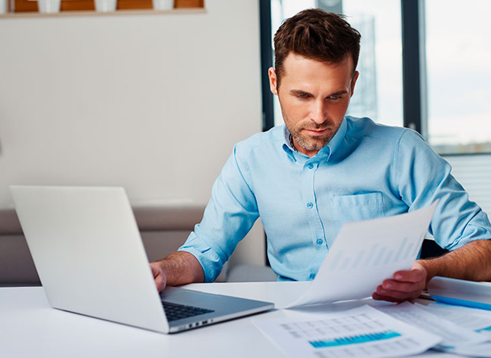 Imagem de um homem branco com camisa social azul sentado numa mesa em frente ao notebook e com um papel na mão.