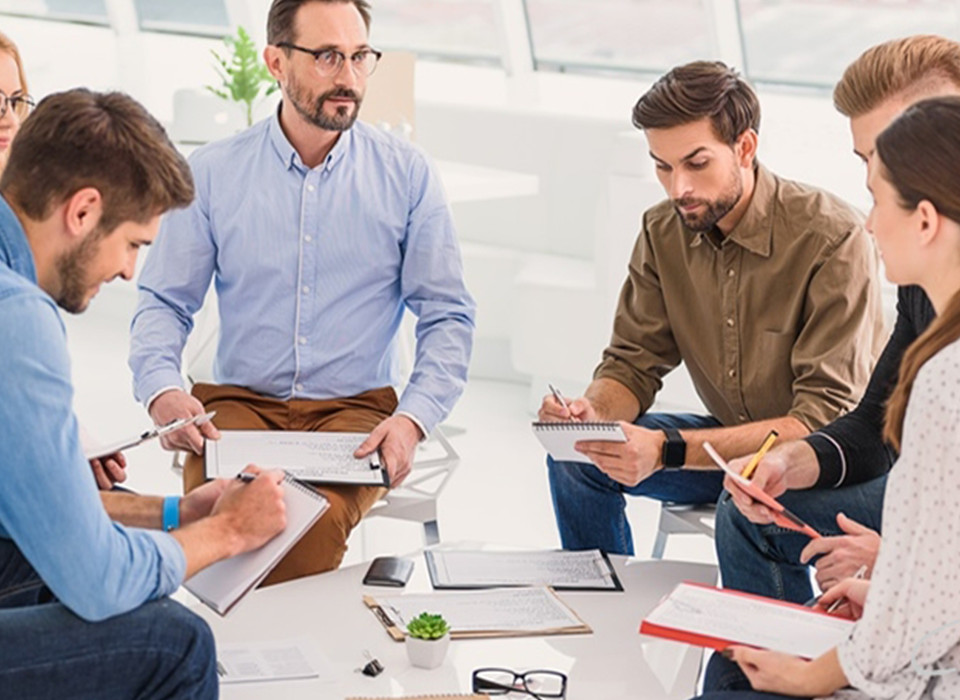 Imagem de um grupo de homens em uma mesa conversando e com cadernos na mão fazendo anotações.