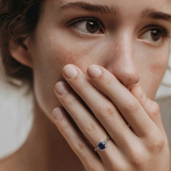 Close-up of woman covering her mouth, wearing a bespoke sapphire engagement ring with diamond halo on her hand