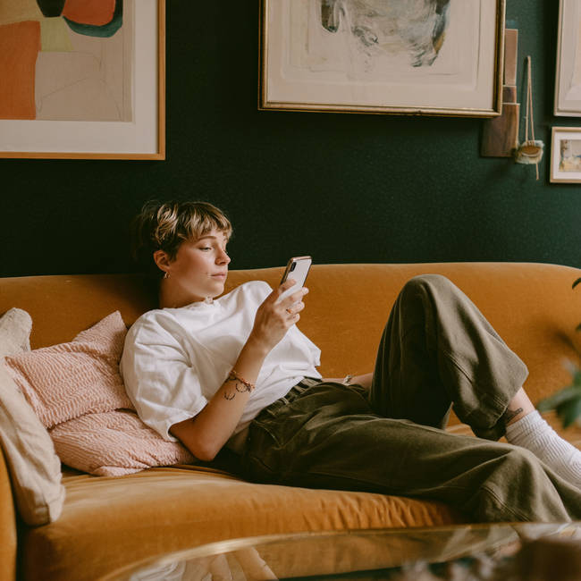Woman relaxing on a sofa, browsing her phone for bespoke engagement ring inspiration in a cosy living room.