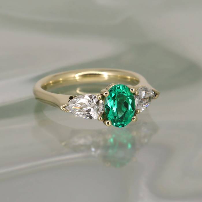 Gold ring with oval green emerald center stone flanked by pear-shaped white diamonds, photographed on a softly reflective pale surface